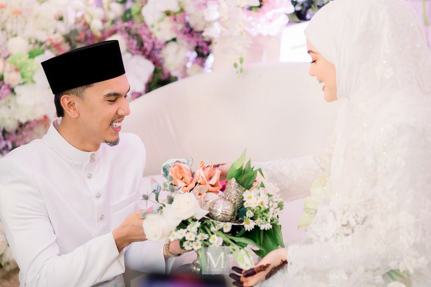 Malaysian groom in white baju melayu presenting a bouquet of pink roses to his bride at their wedding ceremony.