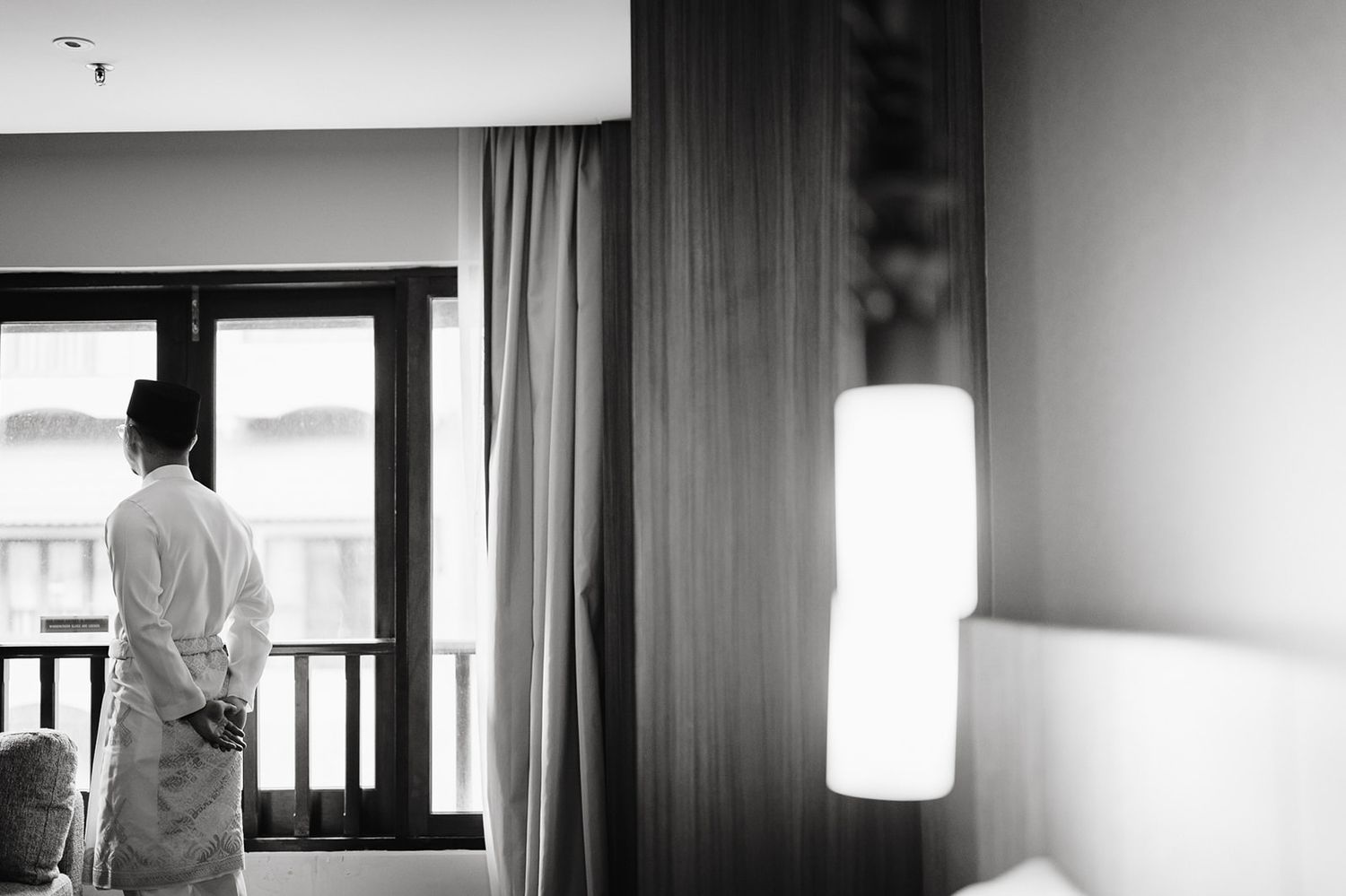 Groom in white robe stands by window in hotel room on wedding morning.