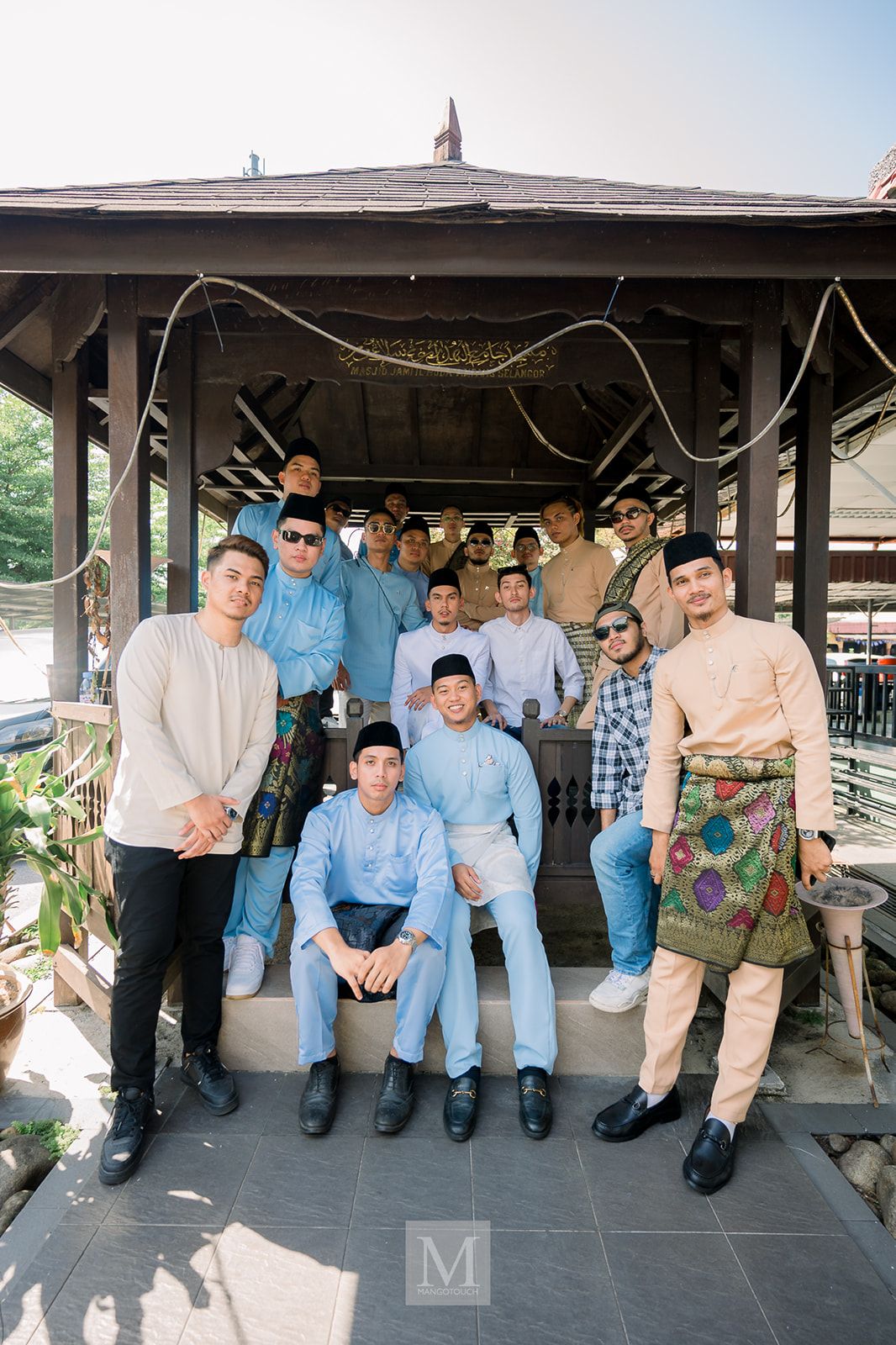 Group of young men in traditional Malay baju melayu posing together under a wooden gazebo at a wedding.