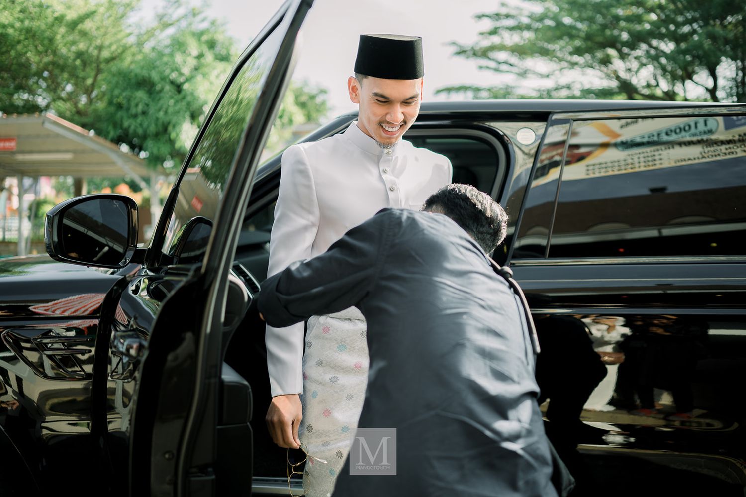 Groom in white baju melayu and black songkok stepping out of a black car assisted by another person.