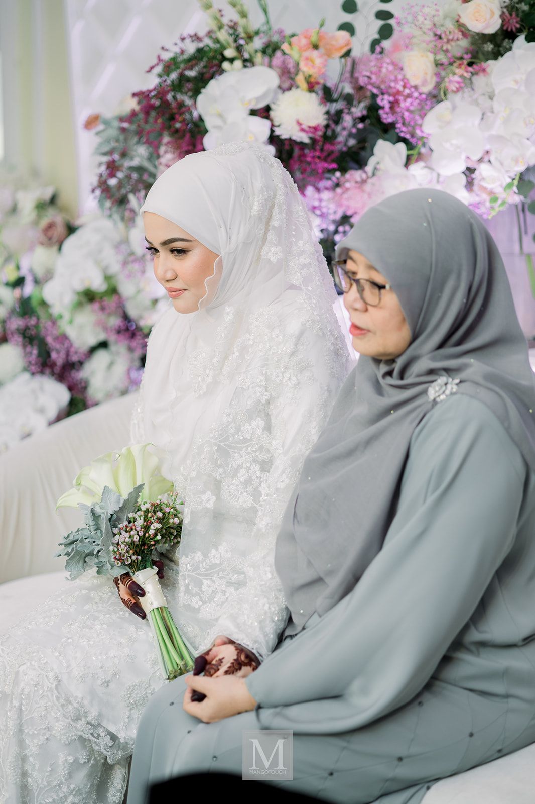 Malaysian bride in white lace gown and hijab holding a bouquet seated beside a woman in grey hijab.