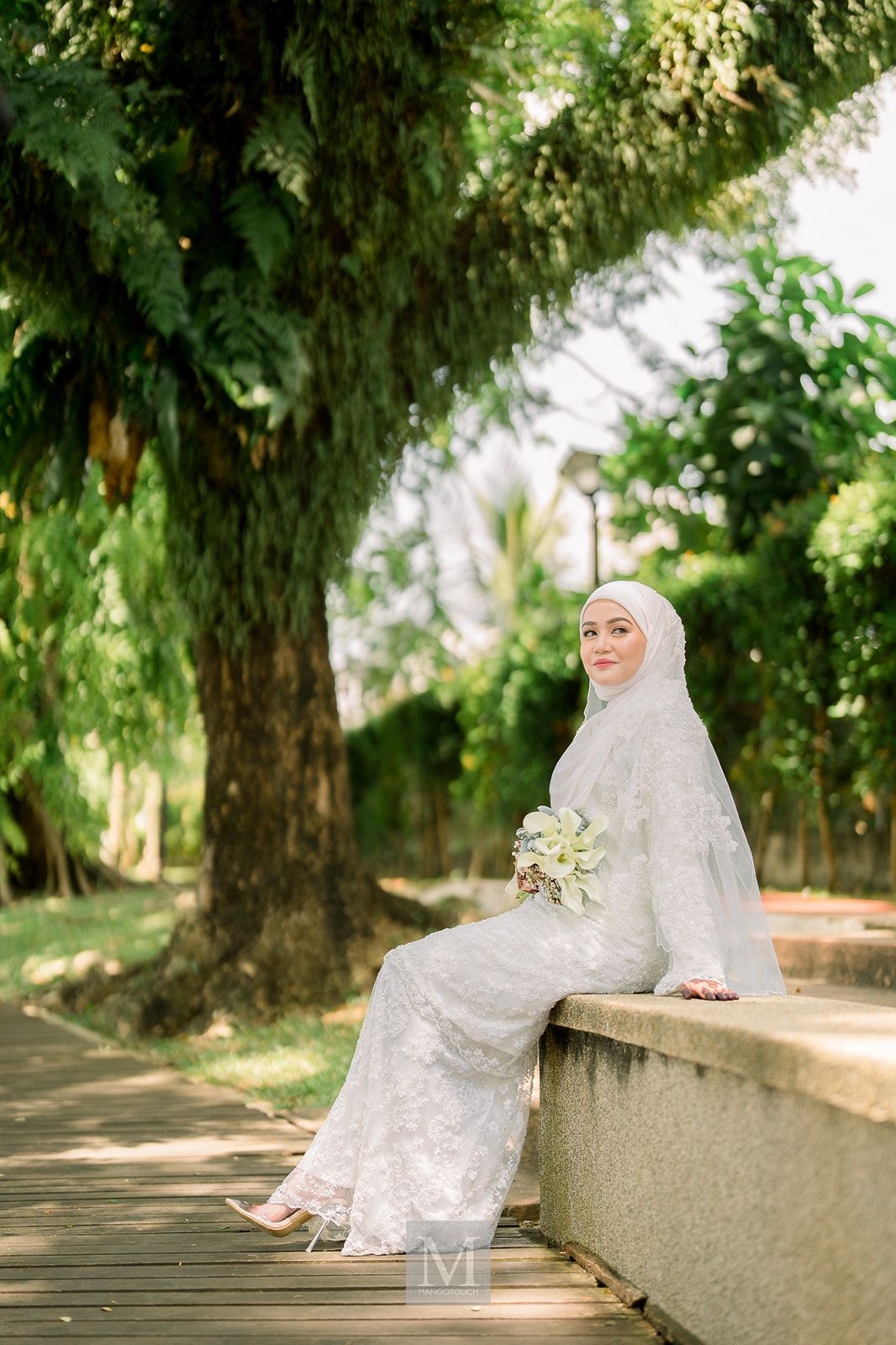 Malaysian bride in white lace wedding dress and hijab sitting outdoors holding a bouquet of white flowers.