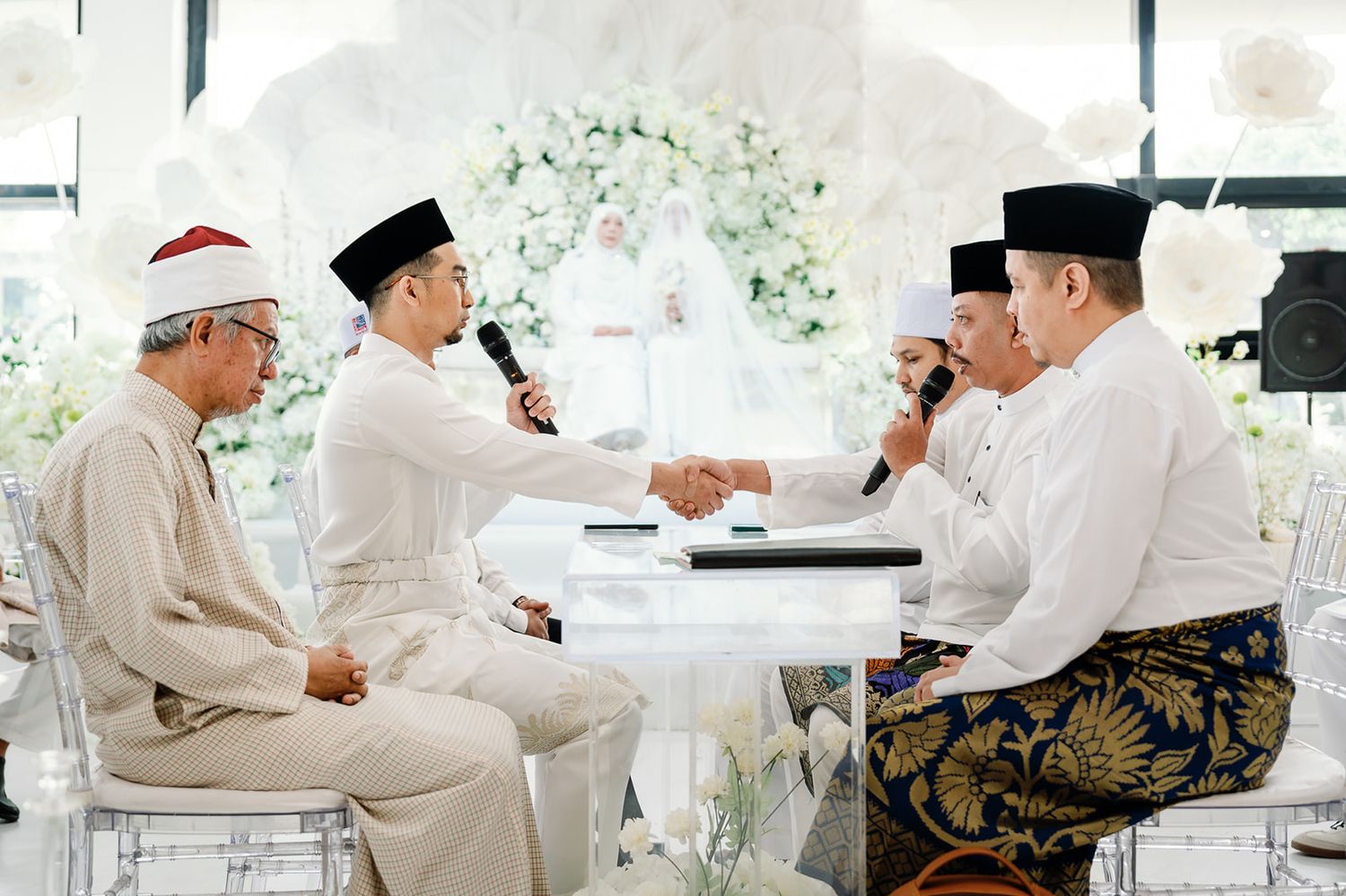 Malaysian groom shaking hands during nikah ceremony at decorated wedding venue.