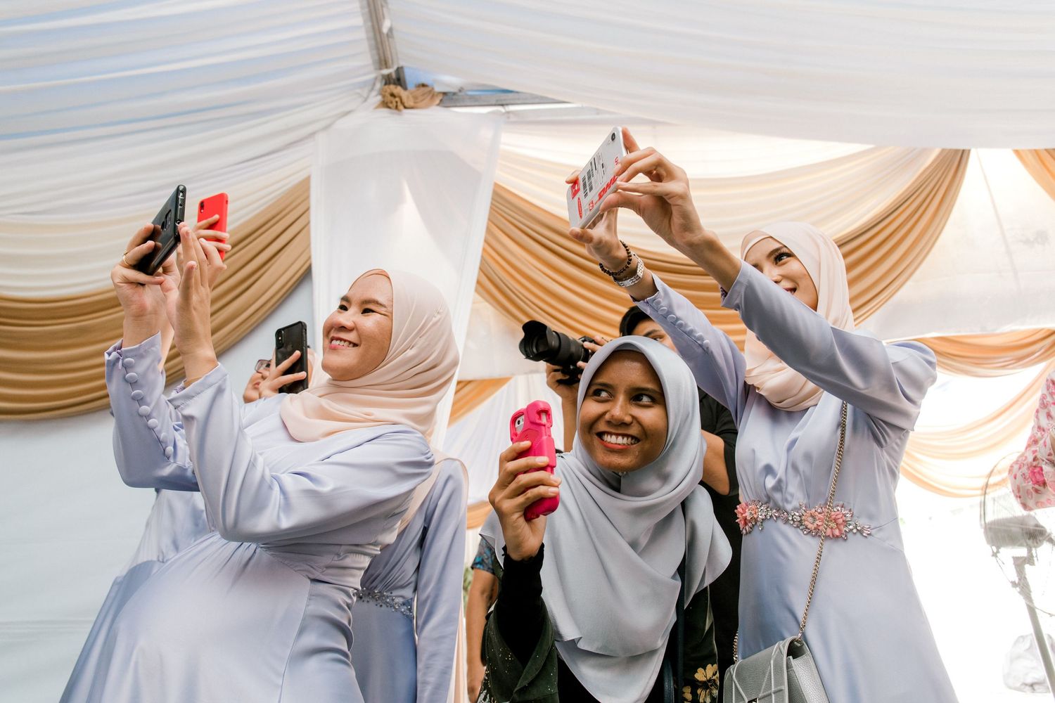 Malaysian  women in hijabs taking selfies with smartphones at a celebration event under decorative draping.