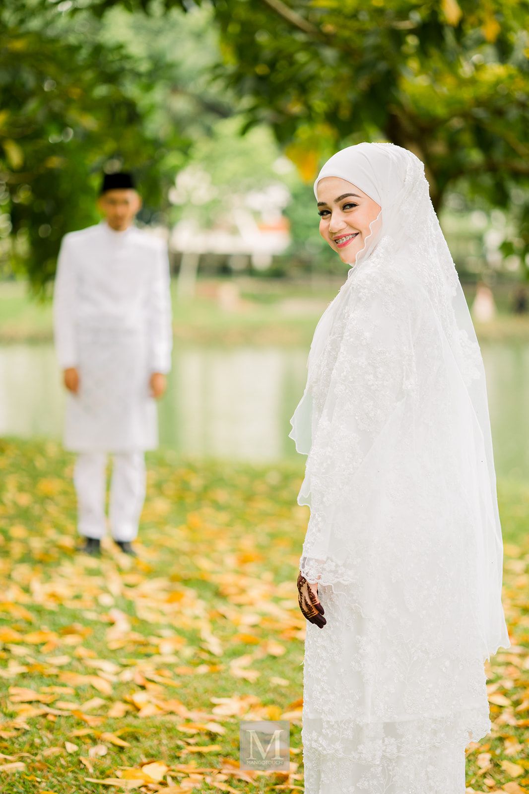Malaysian bride in white hijab and lace wedding dress smiling outdoors with groom in background.