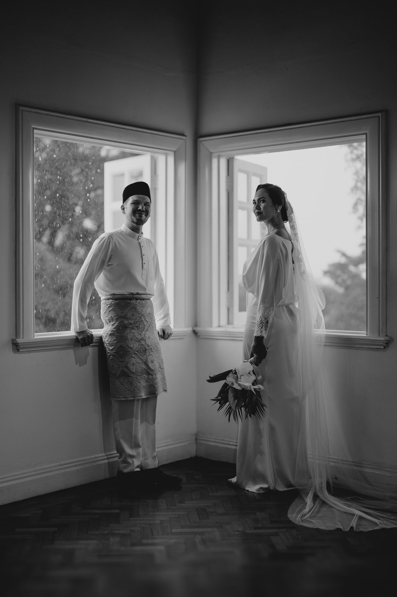 Black and white portrait of Malaysian newlyweds in traditional attire standing by corner windows.