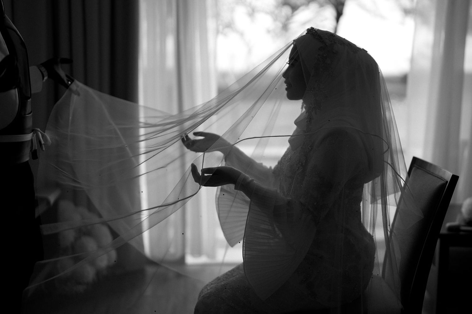 Black and white silhouette of bride in veil sitting by window holding bouquet.