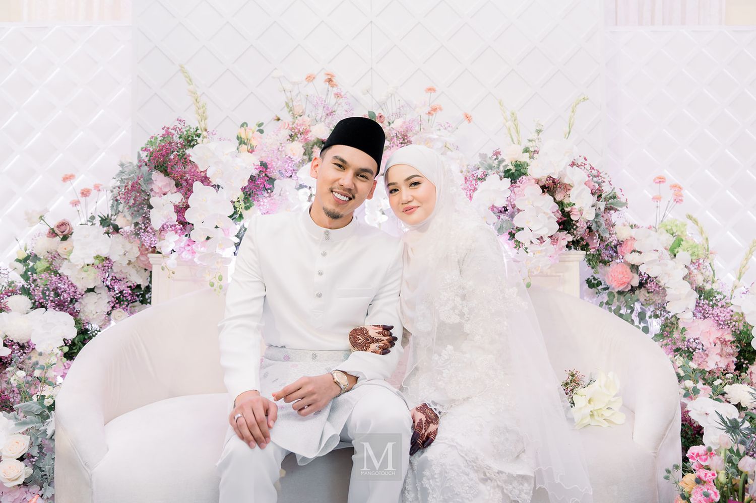 Malaysian bride and groom in white traditional attire posing together on a floral decorated wedding dais.