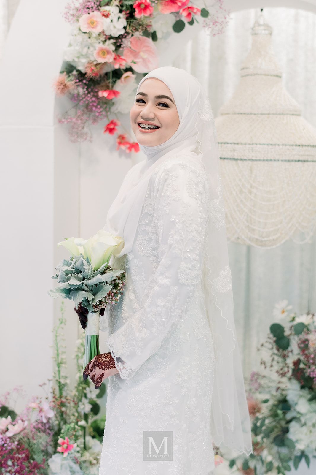 Smiling Malaysian bride in white lace dress and hijab holding a bouquet surrounded by floral decorations.