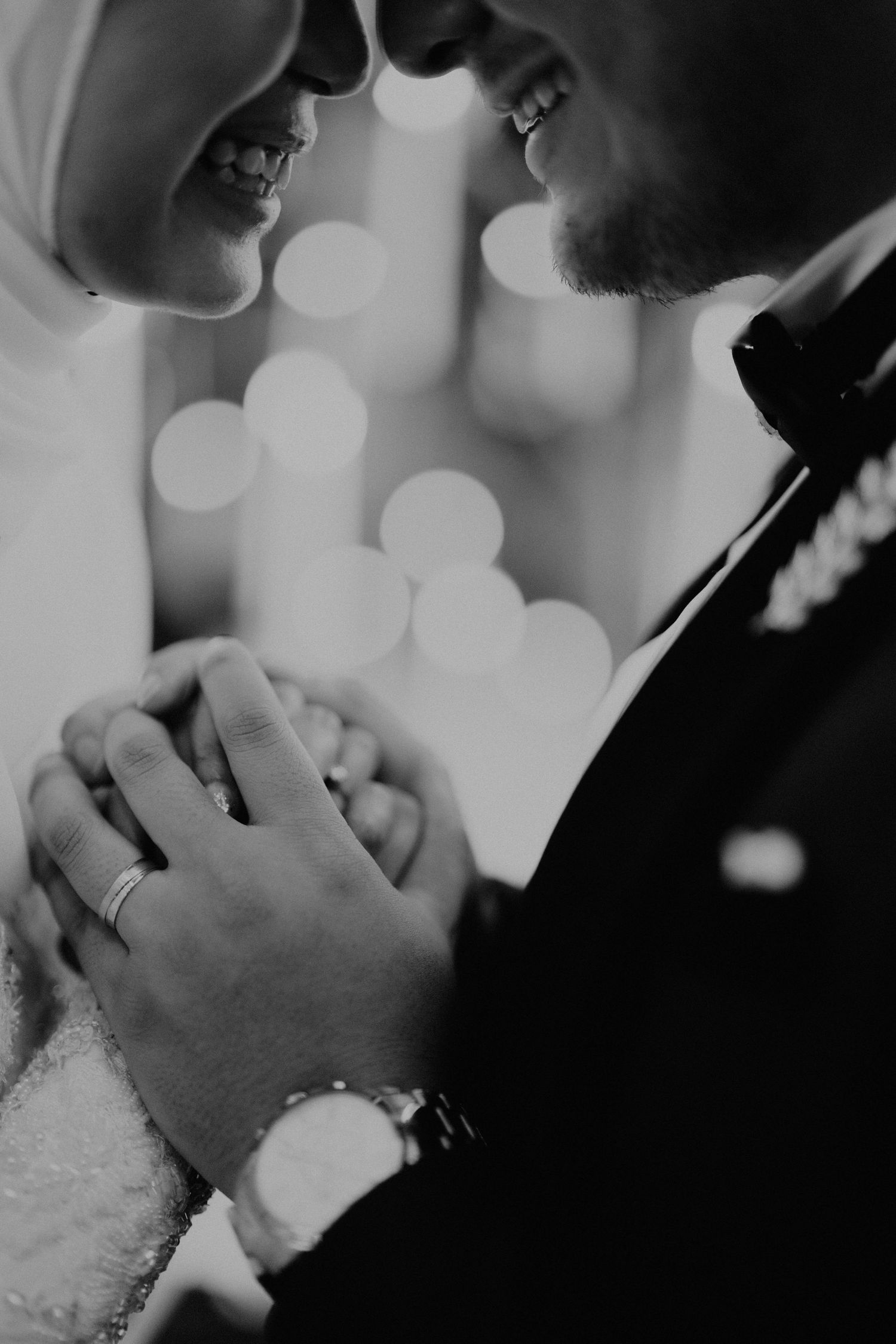 Black and white close-up of newlyweds holding hands with bokeh lights, bride wearing hijab.