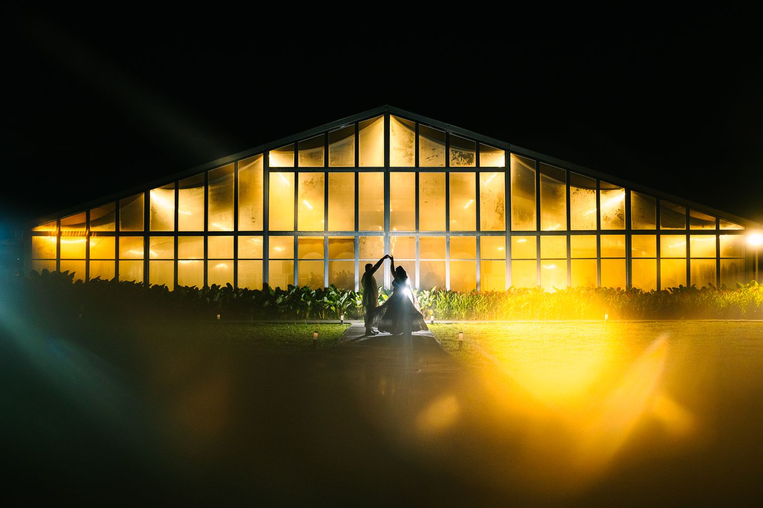 Silhouetted couple posing in front of a glowing greenhouse wedding venue at night.