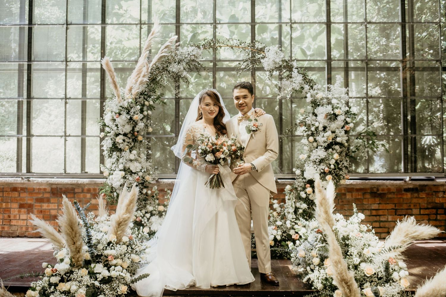 Smiling bride and groom pose under a floral arch with pampas grass in an industrial greenhouse venue.