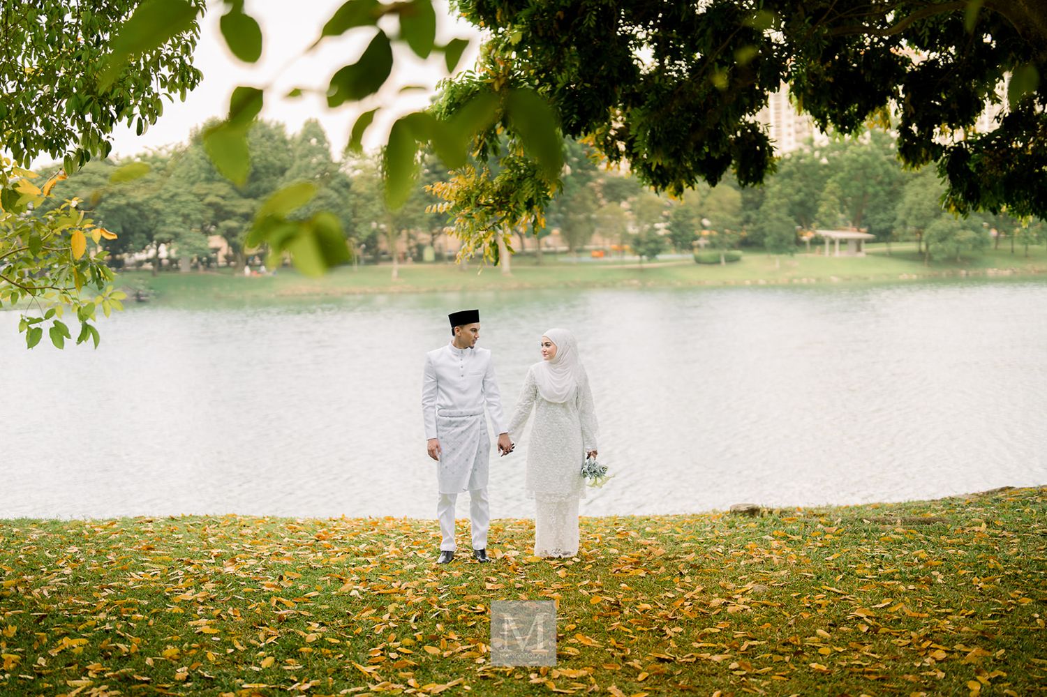 Malaysian  bride and groom in white traditional attire holding hands by a scenic lake surrounded by autumn leaves.