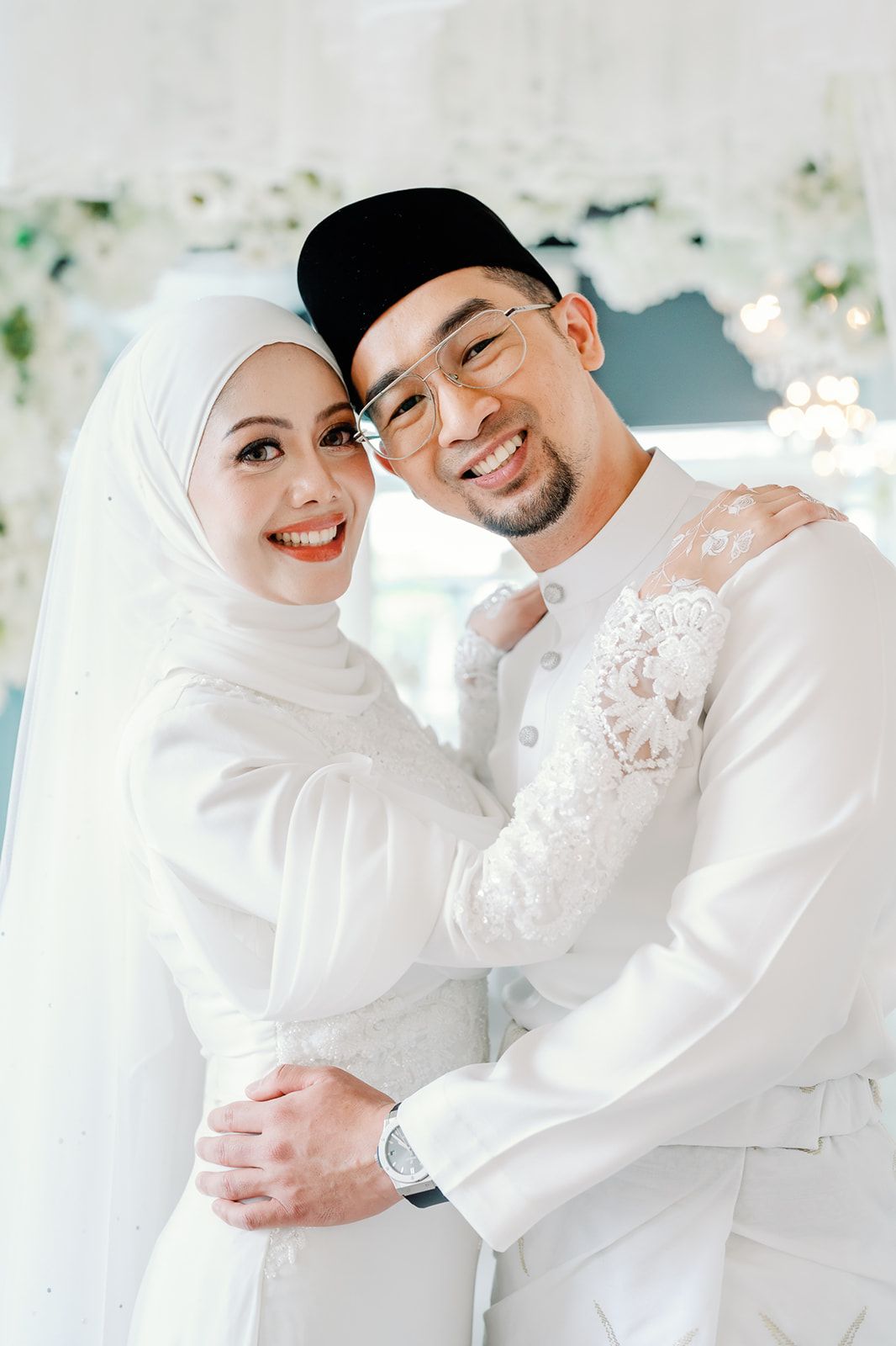 Malaysian couple in matching white wedding outfits embracing and smiling at a beautifully decorated ceremony.