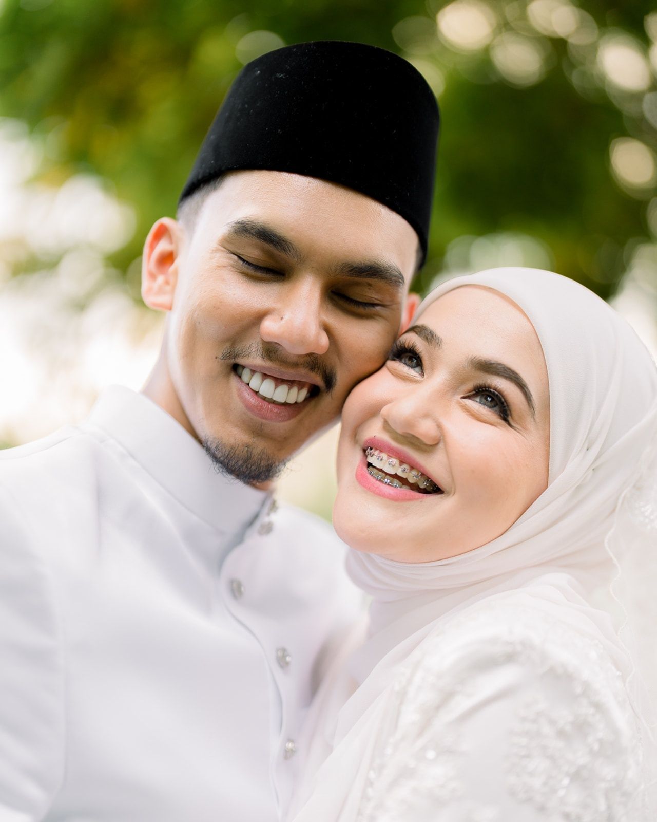 A smiling Malaysian couple in white traditional attire shares a joyful close-up moment outdoors. MK K Clique