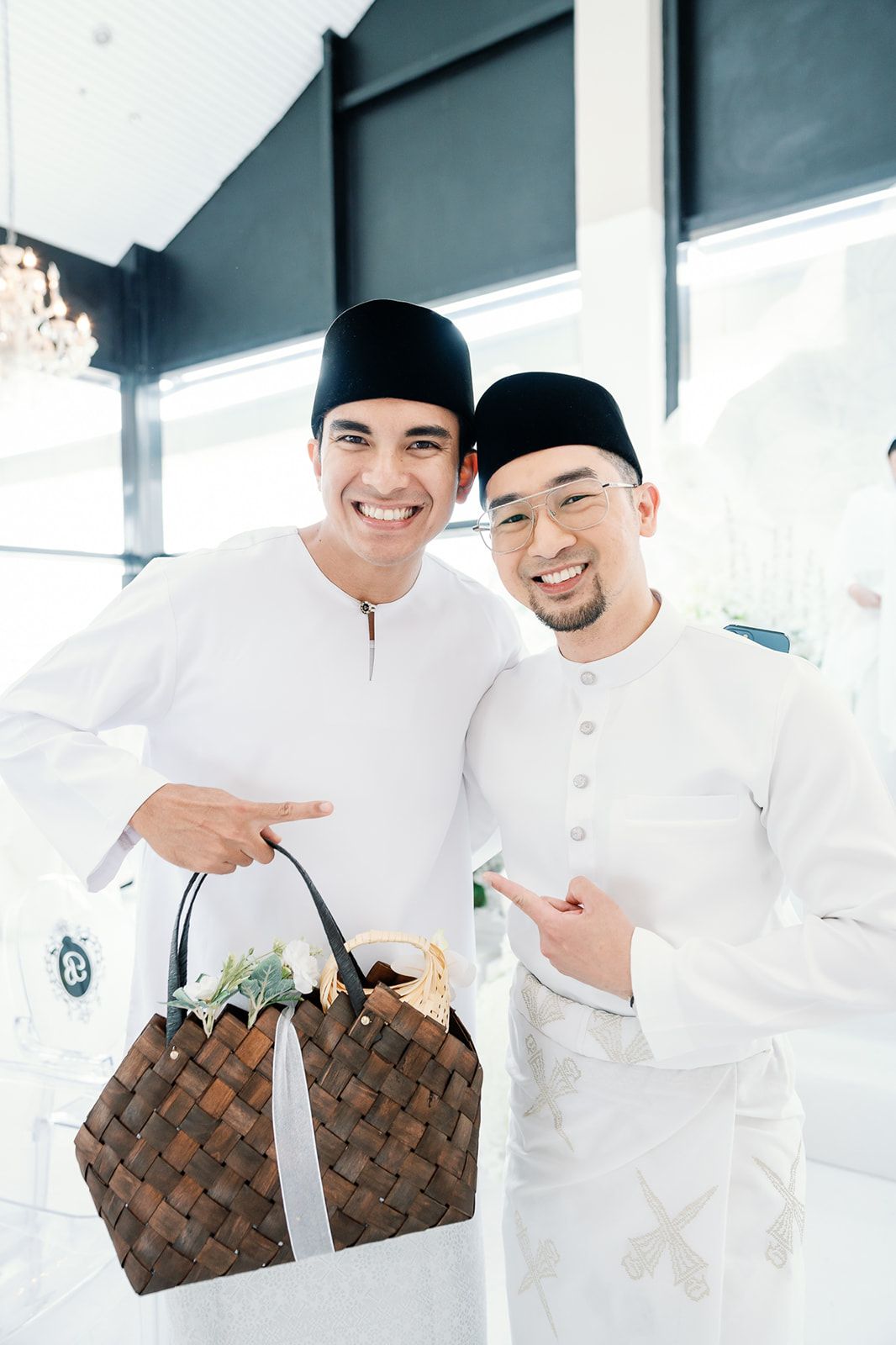 Two Malaysian men in white Baju Melayu and songkok caps posing with a woven basket at a wedding.
