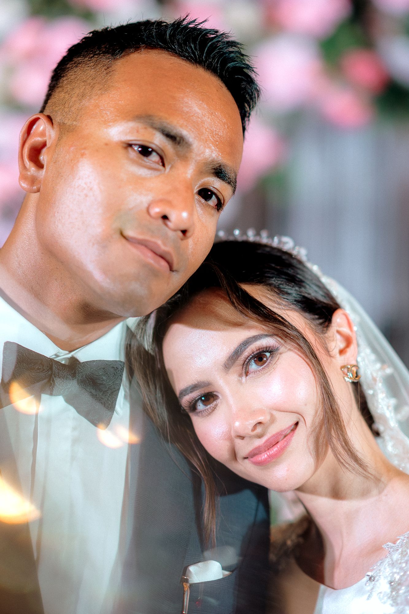 Close-up portrait of newlywed couple, groom in tuxedo and bride in veil with pink floral backdrop.