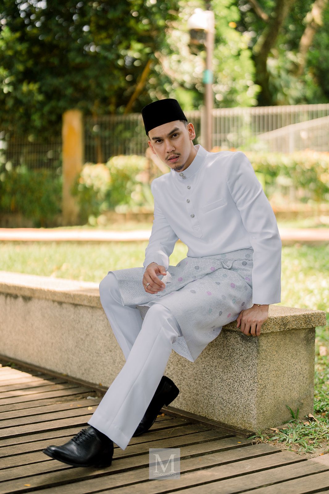 Malaysian groom in white baju melayu and black songkok sitting outdoors in a park setting.