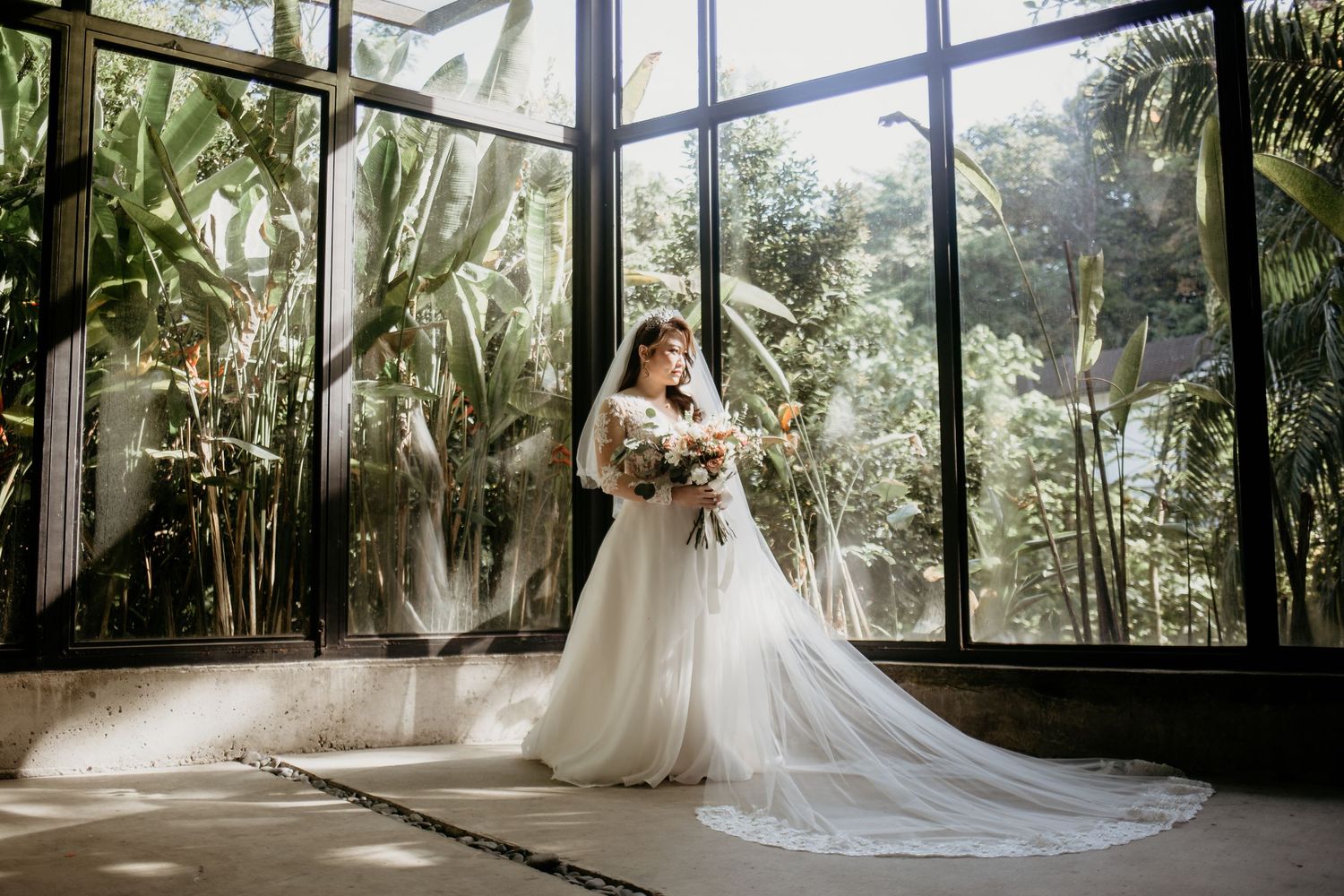 Elegant bride in white gown with long train standing before large greenhouse windows with tropical plants.