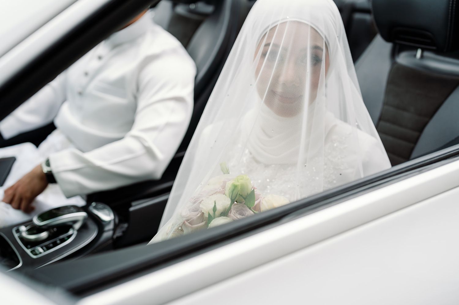 Malaysian bride in white hijab and veil holding bouquet seated in luxury wedding car.