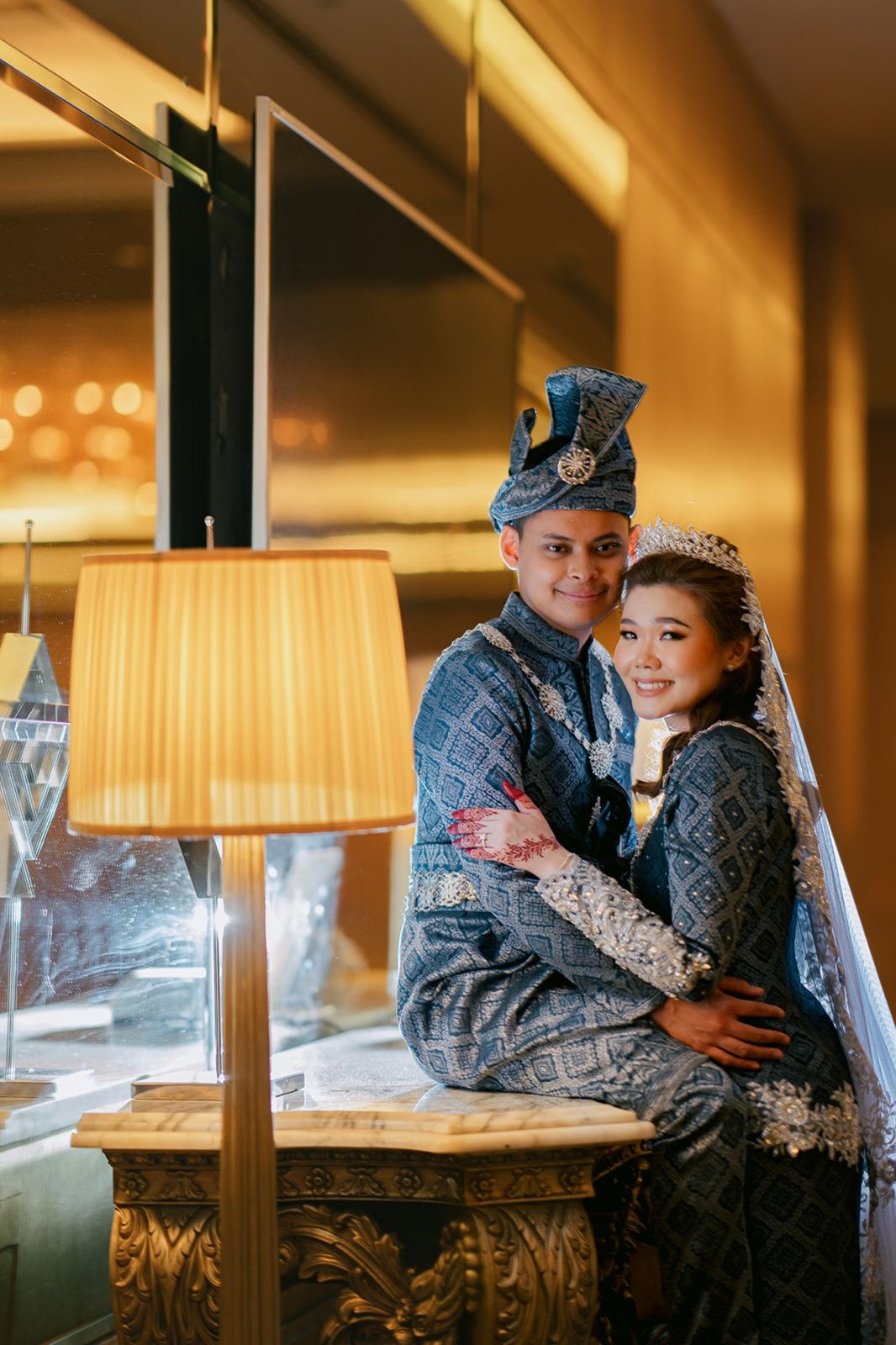 Malay couple in elegant blue traditional wedding attire embracing warmly in a luxurious golden interior setting.
