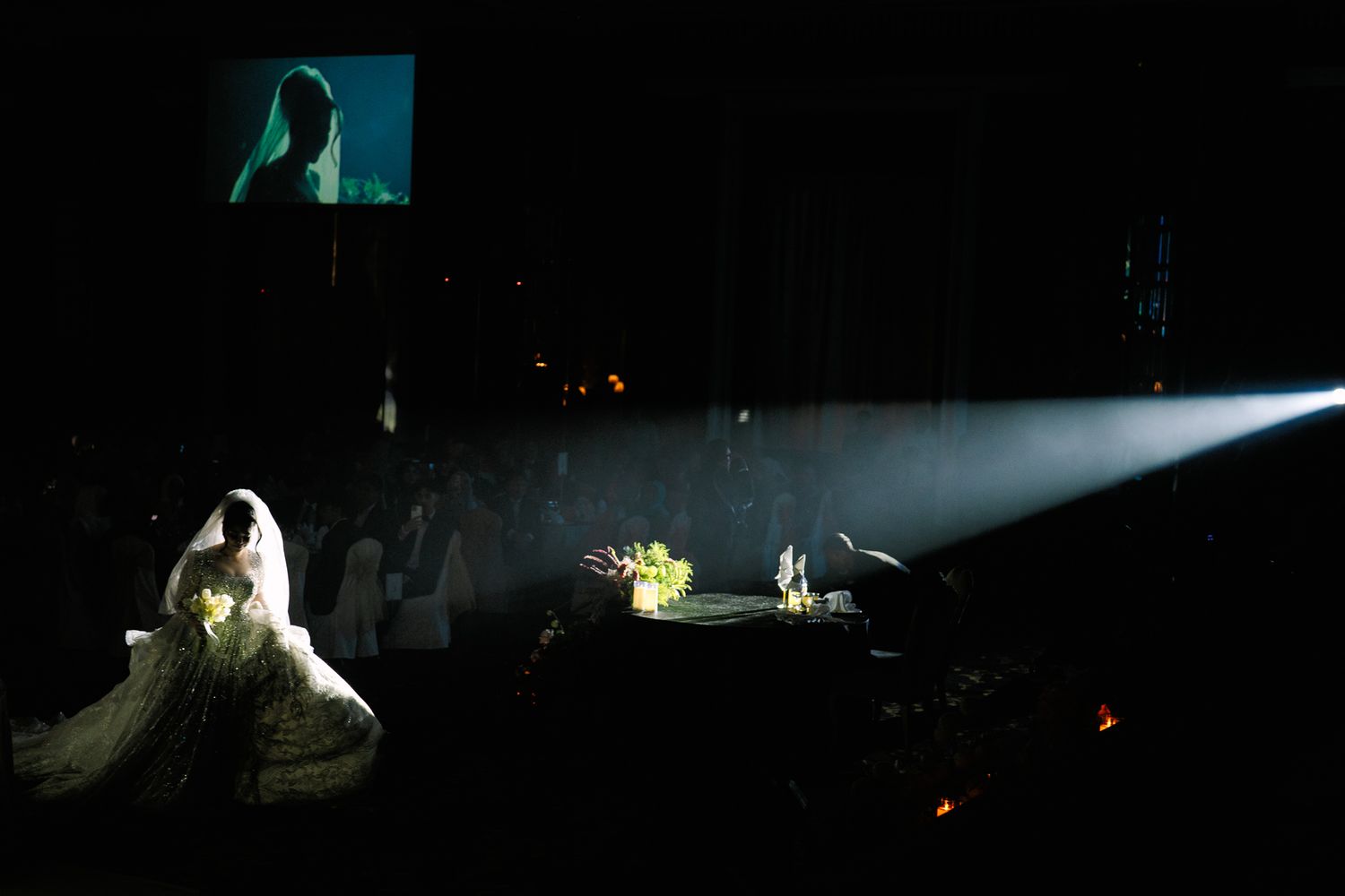 Dramatic wedding reception scene with spotlight on bride in gown and projected screen above stage.