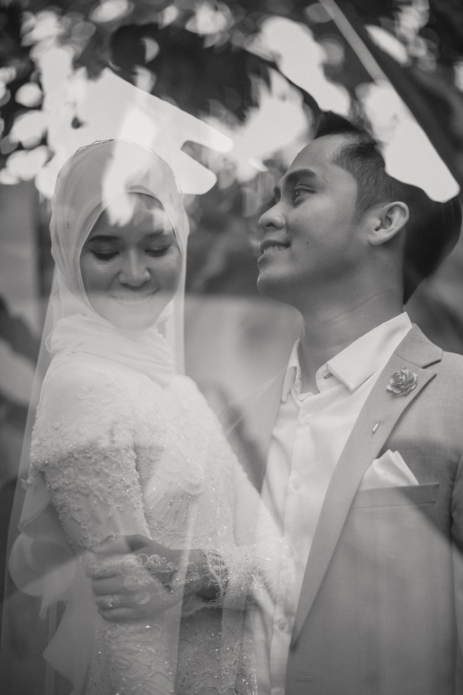 Black and white wedding photo of a smiling Malaysian couple, bride in hijab and lace dress, groom in suit, outdoors.