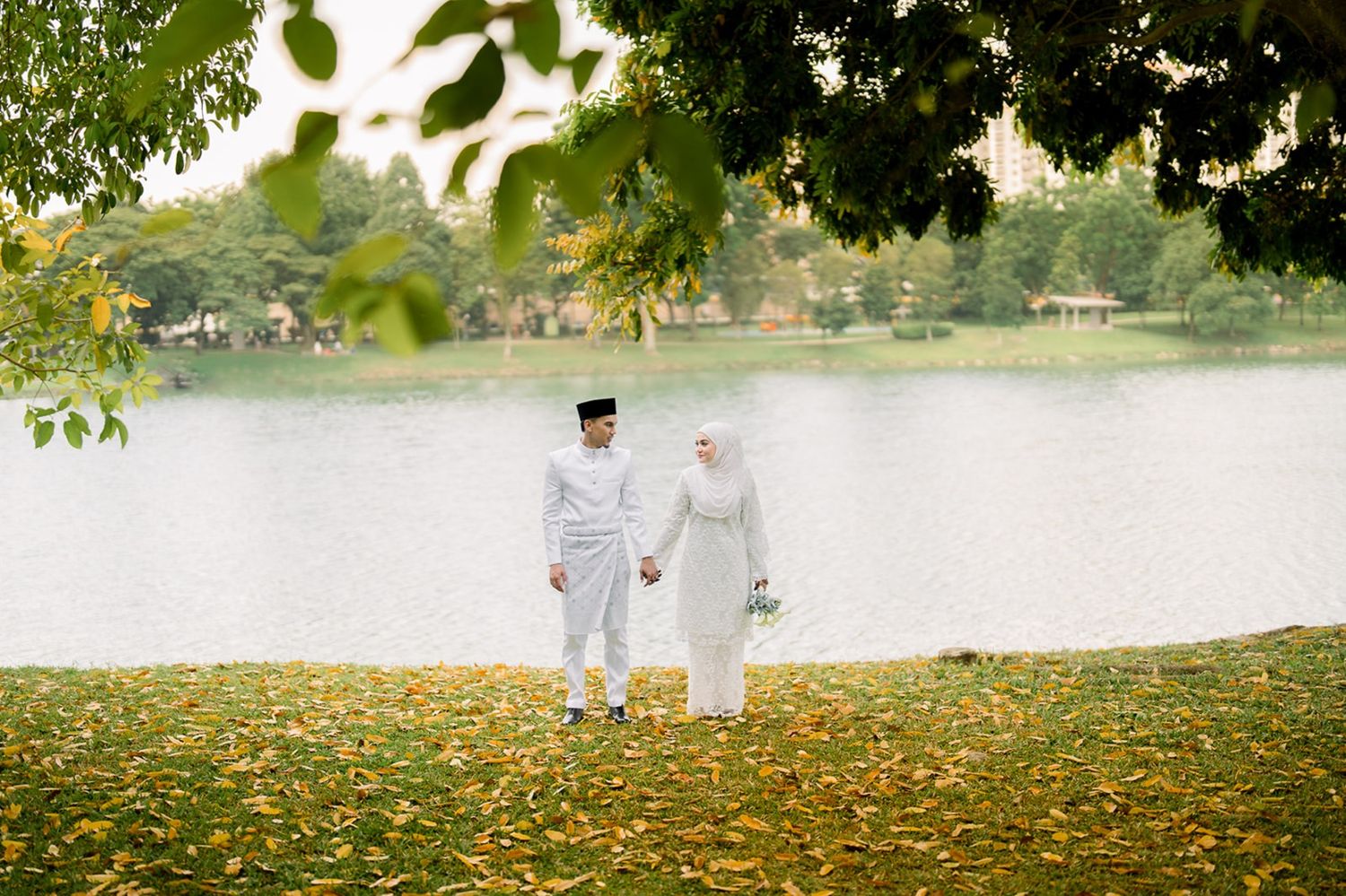 A Malaysian couple in white traditional wedding attire stands hand-in-hand by a peaceful autumn lakeside. MK K Clique