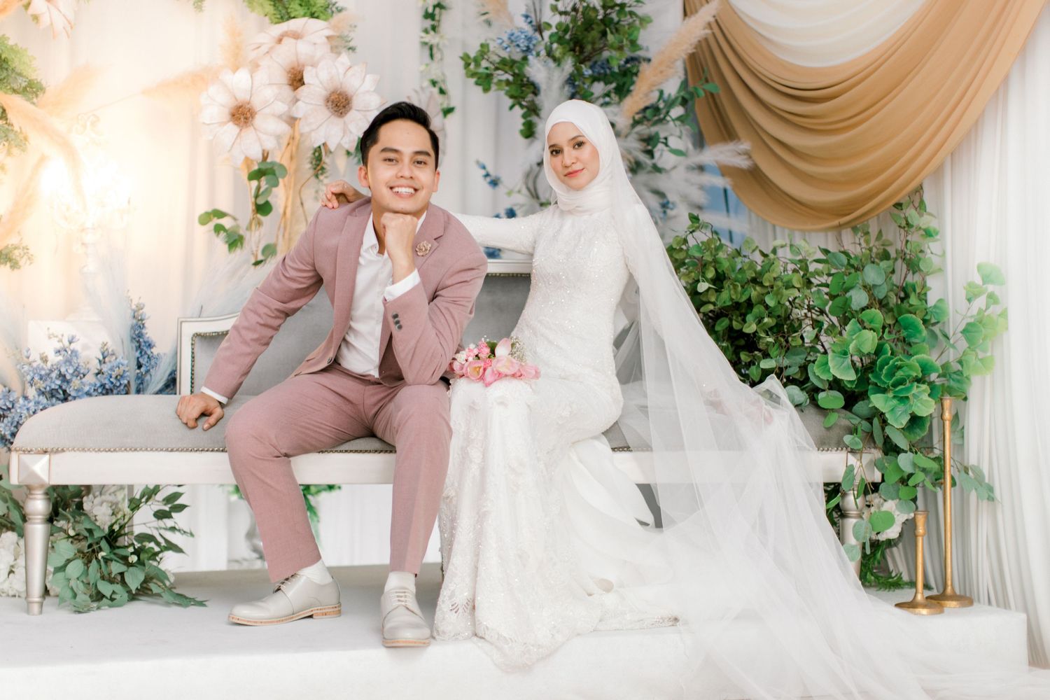 Malaysian couple in wedding attire, groom in pink suit and bride in white hijab gown, posing at decorated reception backdrop.