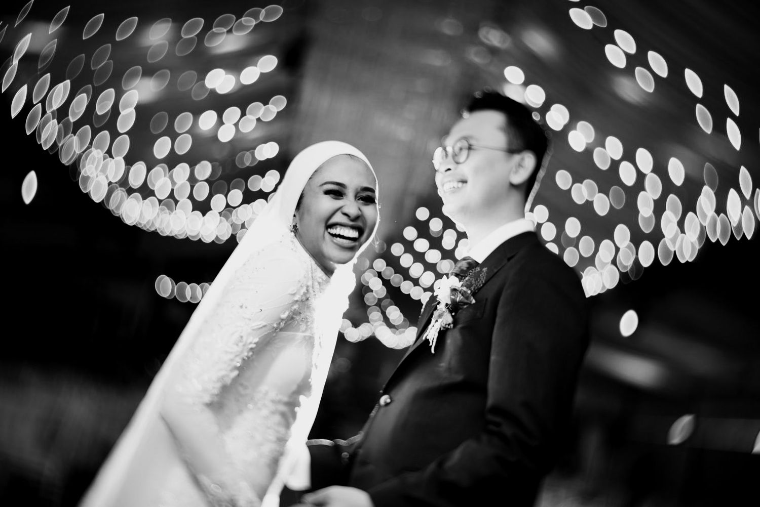 Black and white photo of laughing bride in hijab and groom under bokeh chandelier lights.