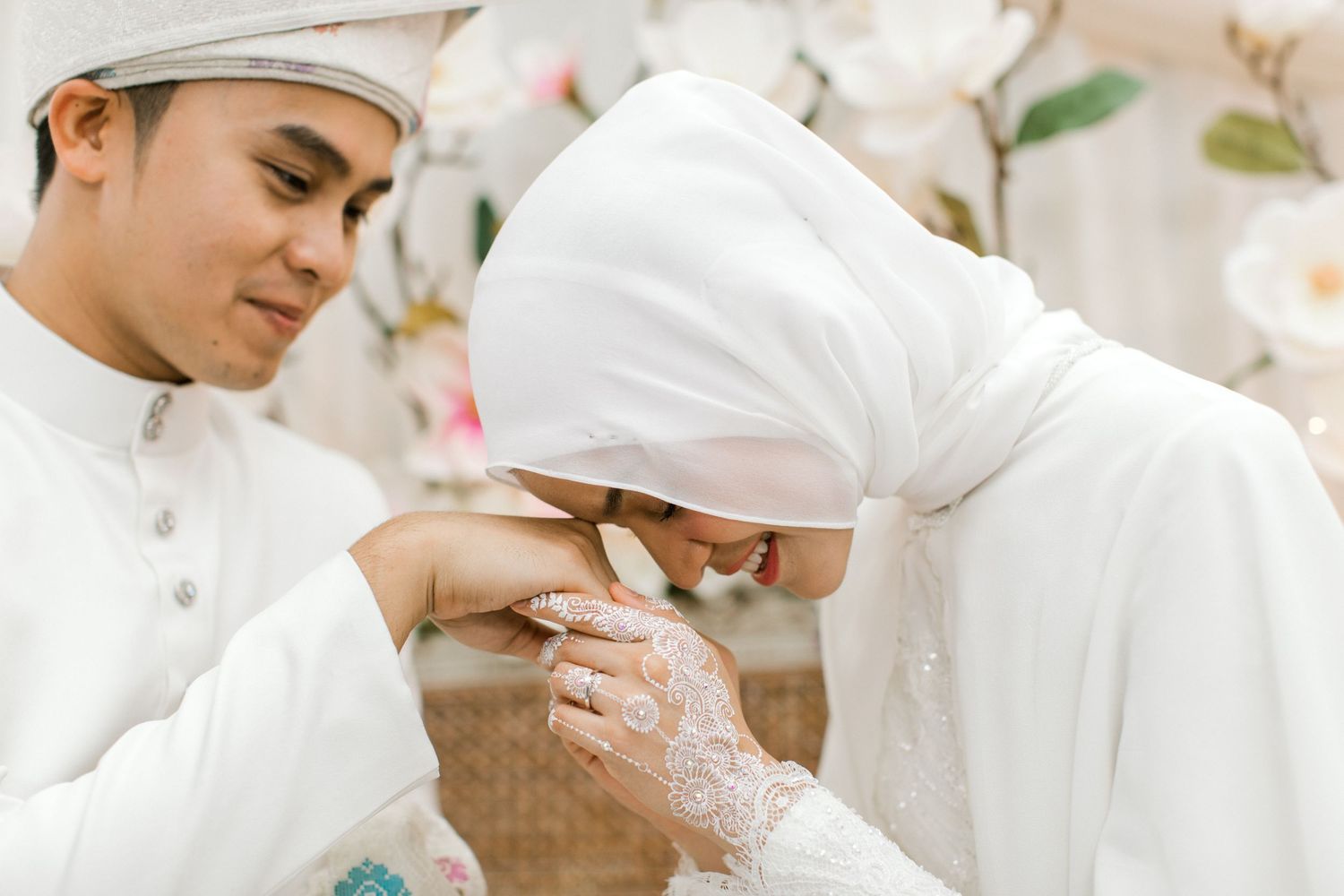 Malaysian groom kissing brides hand adorned with white henna at traditional Malay wedding ceremony.