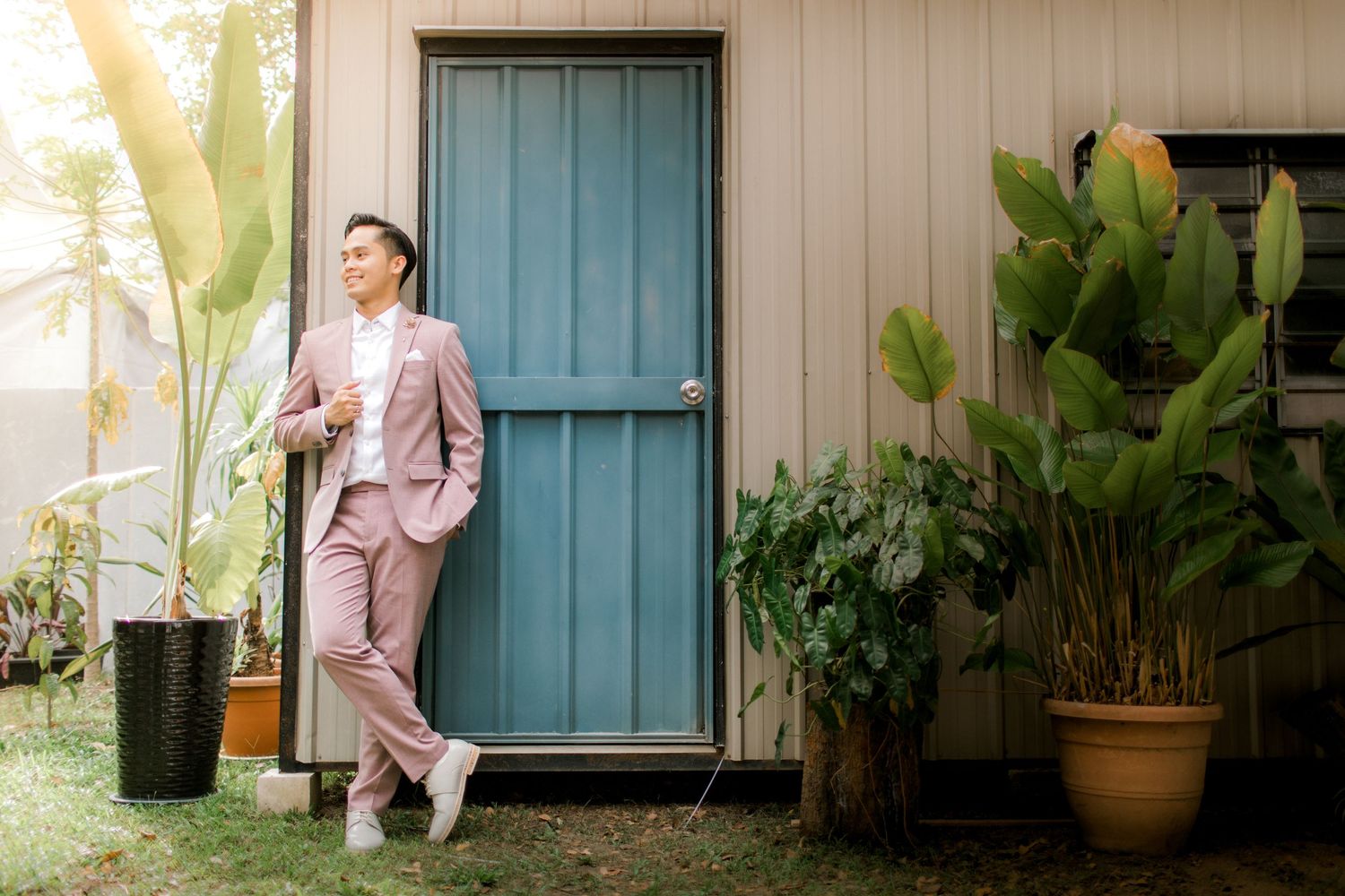 Man in pink suit leaning against blue door surrounded by tropical plants in outdoor setting.