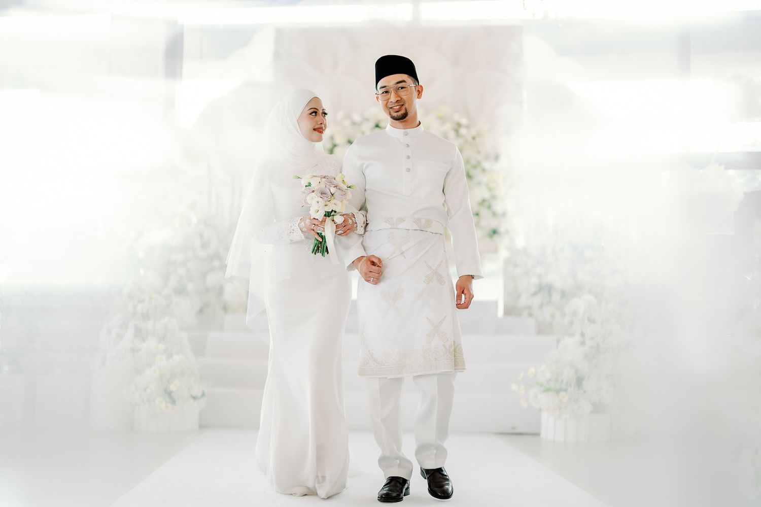 Malaysian bride and groom in matching white traditional attire posing at their wedding ceremony.