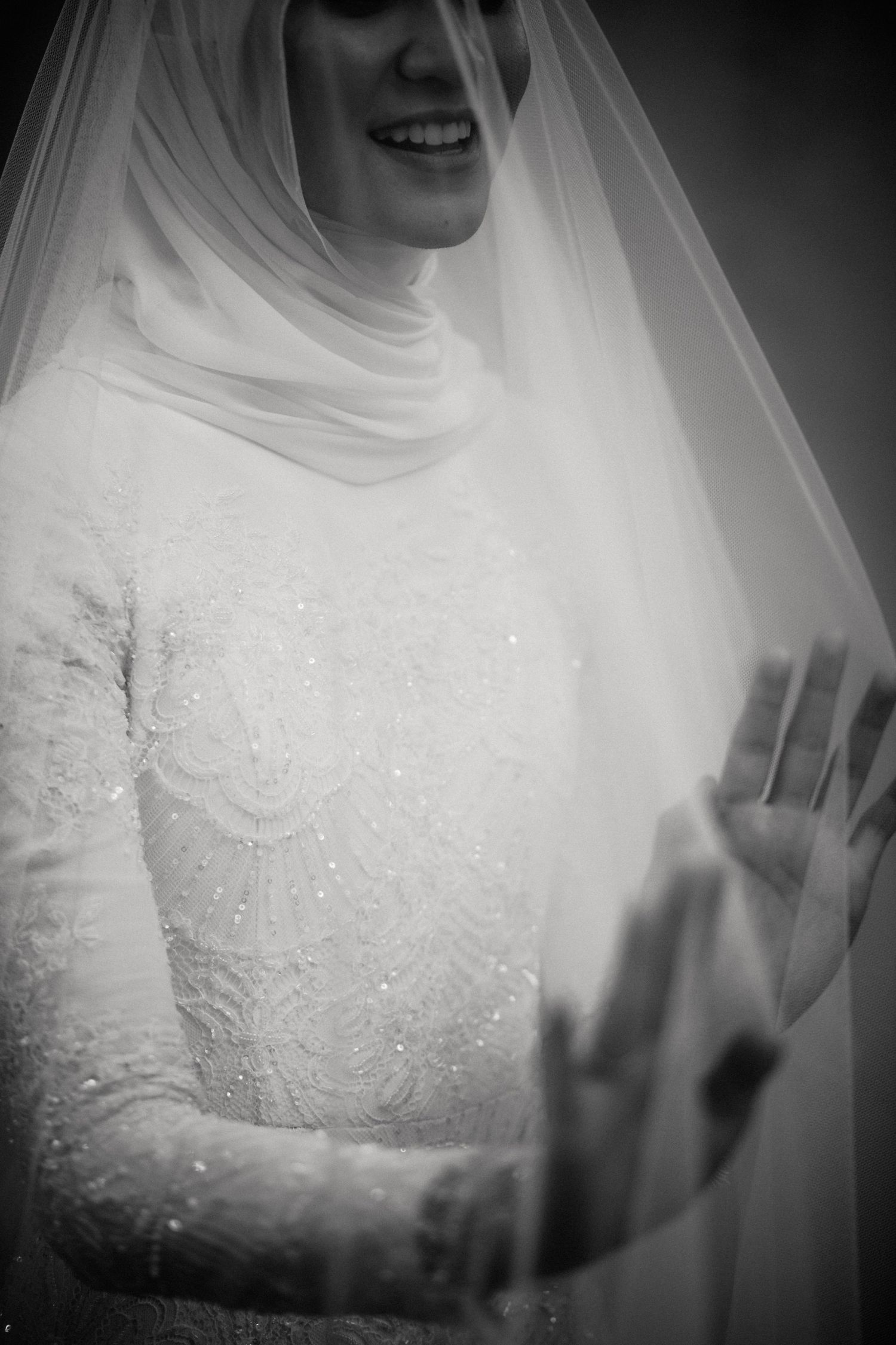 Black and white photo of a smiling Malaysian bride in a white lace gown and hijab, partially covered by a flowing veil.