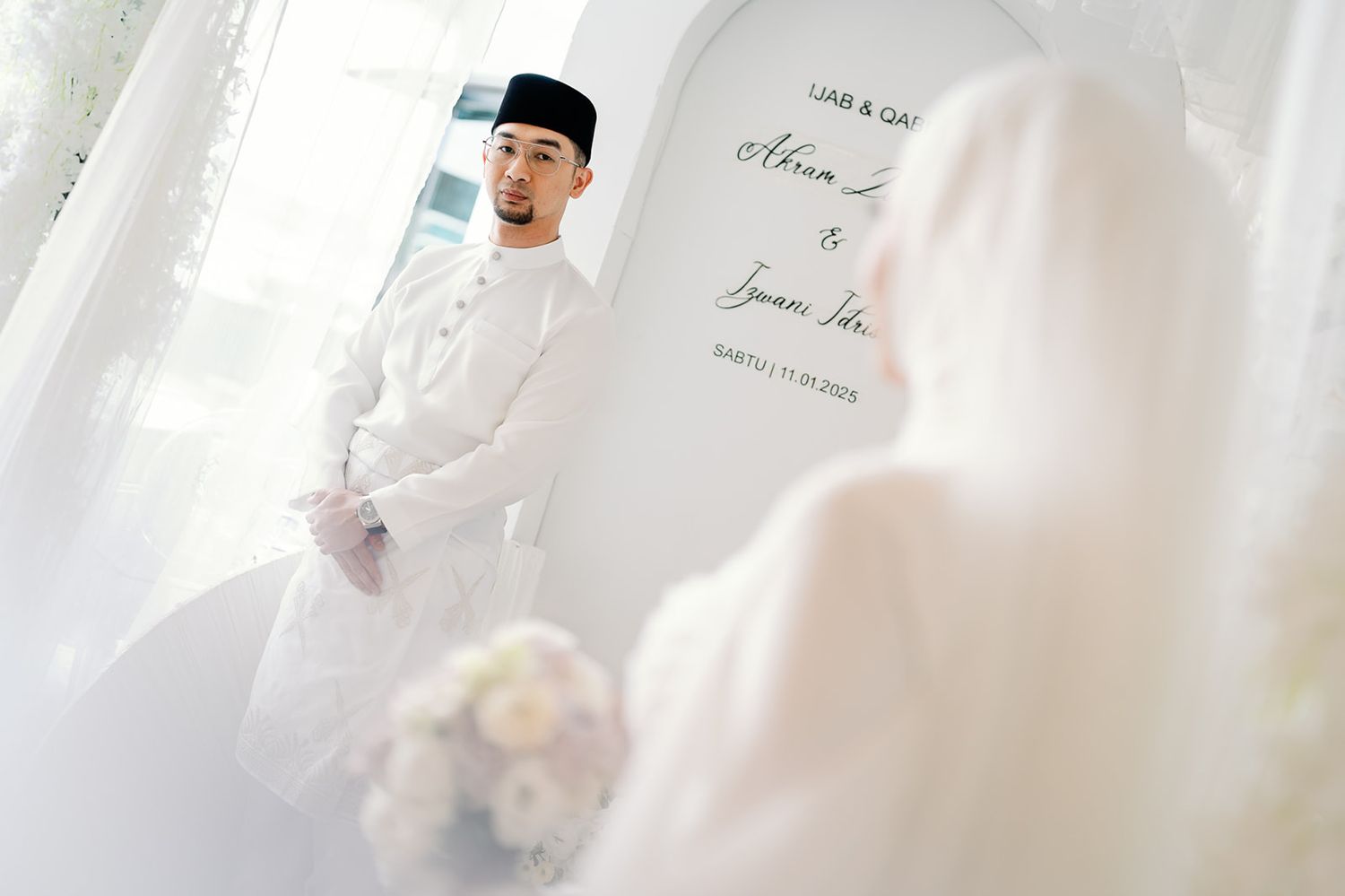 Malaysian groom in white Baju Melayu and songkok gazing at his bride during wedding ceremony.