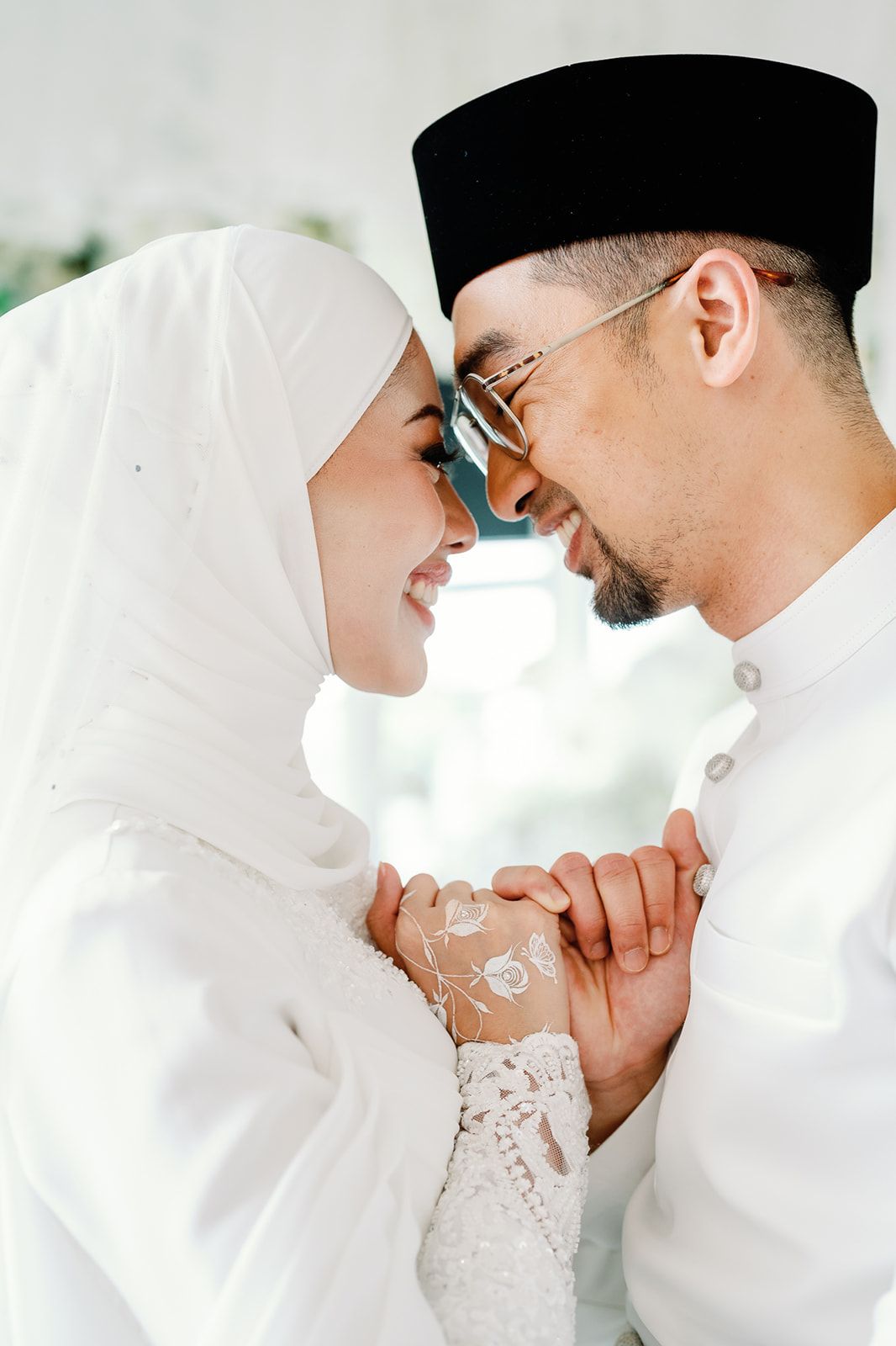 Malaysian couple in white attire sharing a romantic moment, holding hands and touching foreheads on their wedding day.