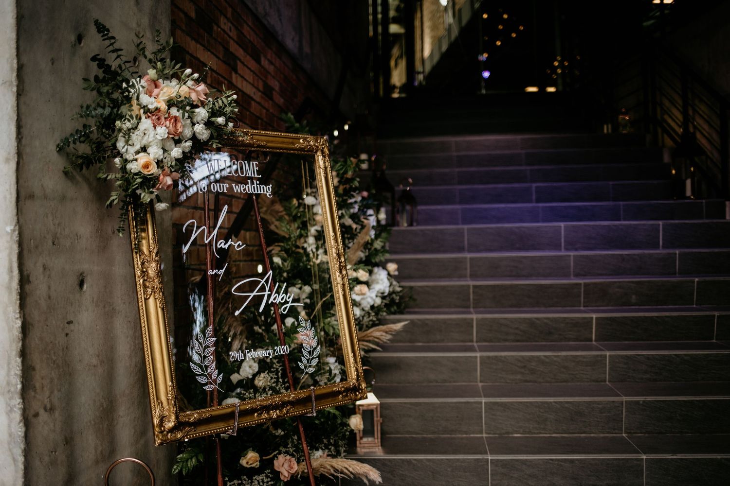 Elegant gold-framed wedding welcome mirror sign adorned with lush floral arrangements beside stone staircase.