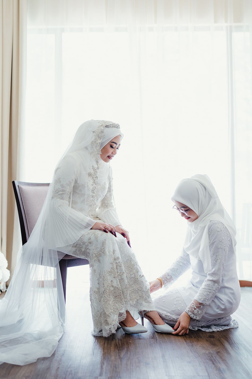 Malaysian bride in white lace kebaya sits as bridesmaid helps put on silver heels before wedding ceremony.