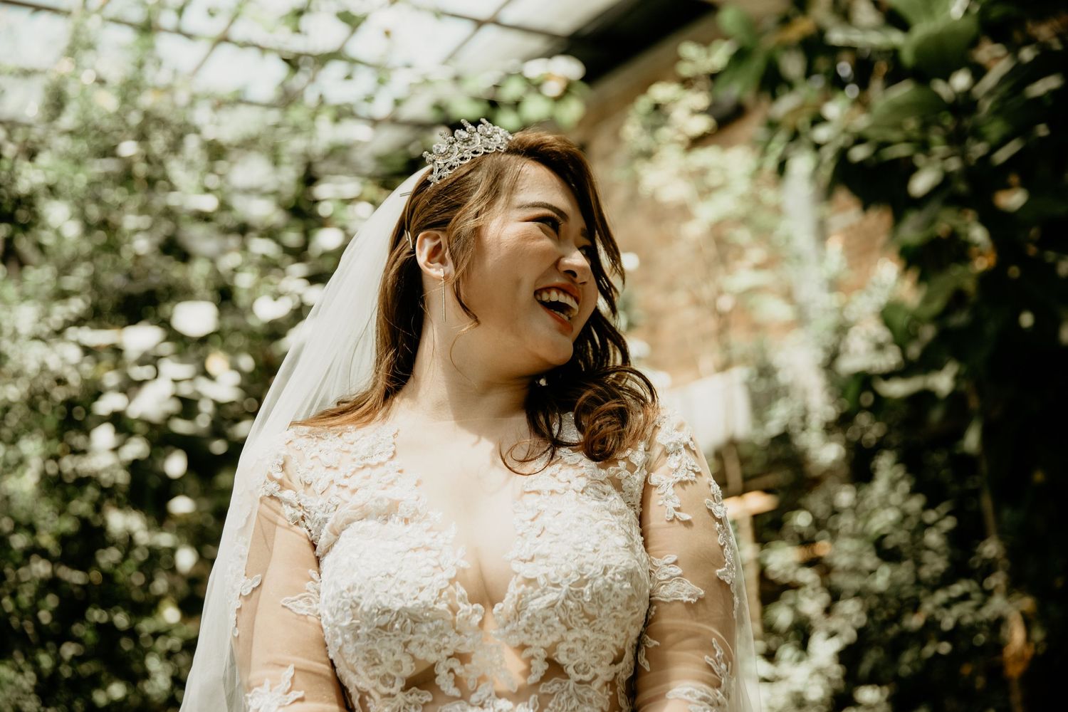 Radiant bride wearing a lace gown and tiara smiles joyfully surrounded by lush greenery.