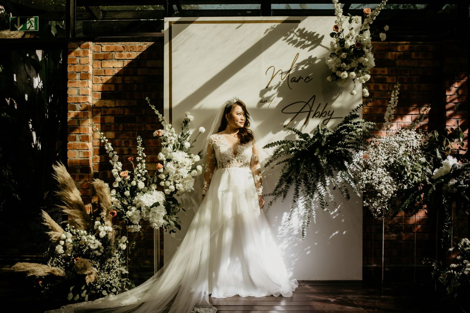 Bride in flowing white gown posing before floral arch with ferns and white blooms in greenhouse venue.