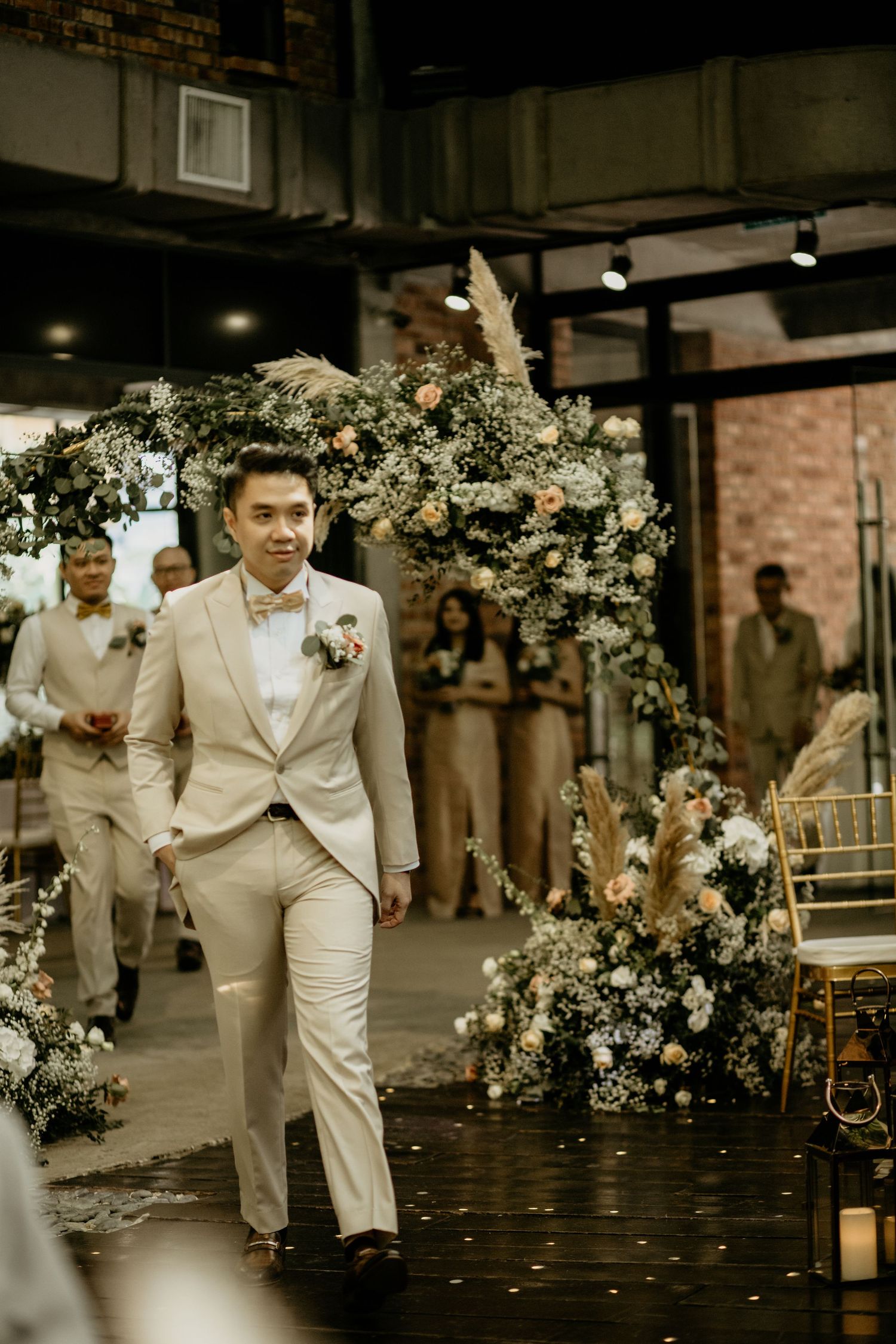 Dapper groom in beige suit with bow tie and boutonniere walks down floral-decorated wedding aisle.