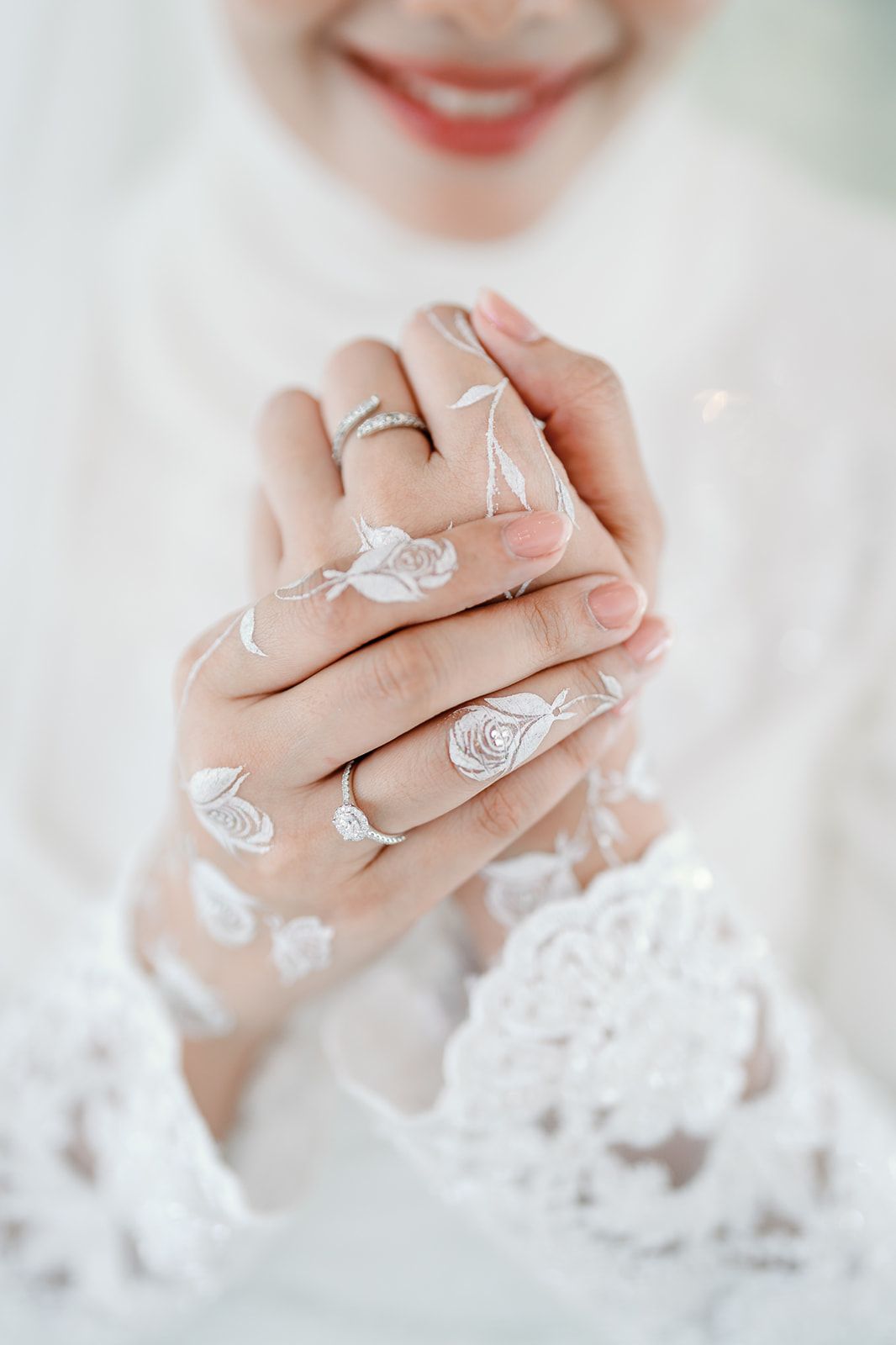 Close-up of a bride's hands adorned with silver rings and white floral henna art, wearing elegant white lace dress.