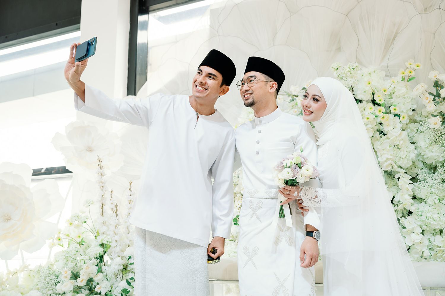 Malaysian wedding couple taking a selfie with a guest in traditional white Baju Melayu attire.