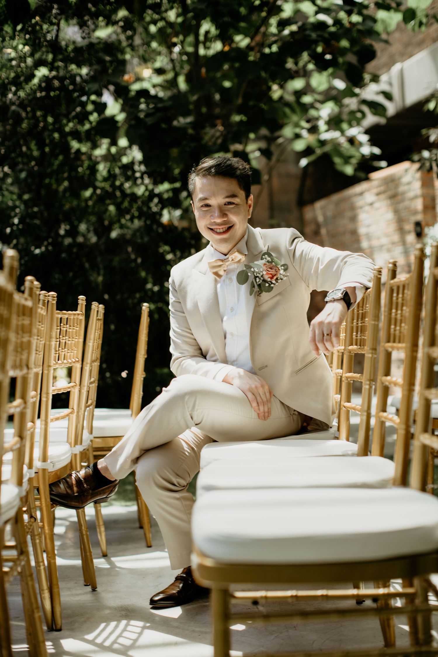 Dapper groom in a beige suit sits relaxed among gold chiavari chairs at an outdoor wedding venue.
