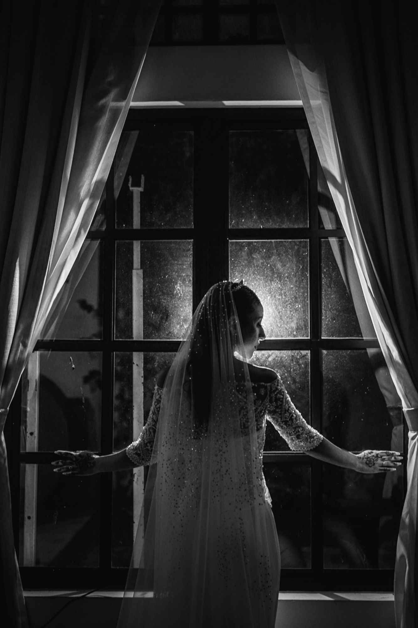 Black and white photo of a bride in lace gown standing at a large window with curtains.