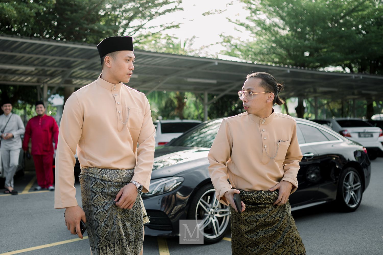 Two men in matching beige baju melayu traditional attire conversing outdoors near a black car.