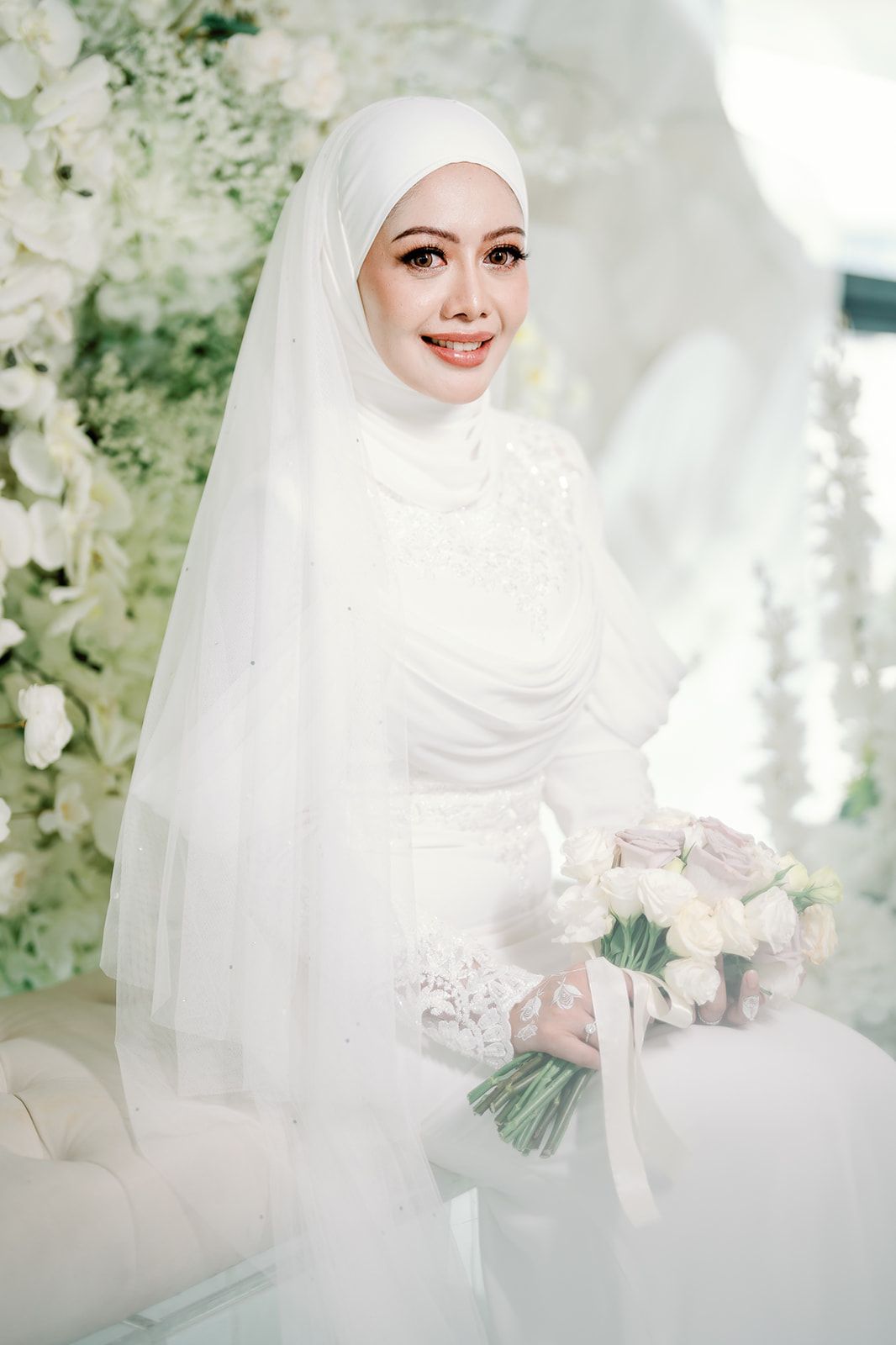 Beautiful Malaysian bride in white hijab and lace gown holding pastel bouquet surrounded by white floral decor.