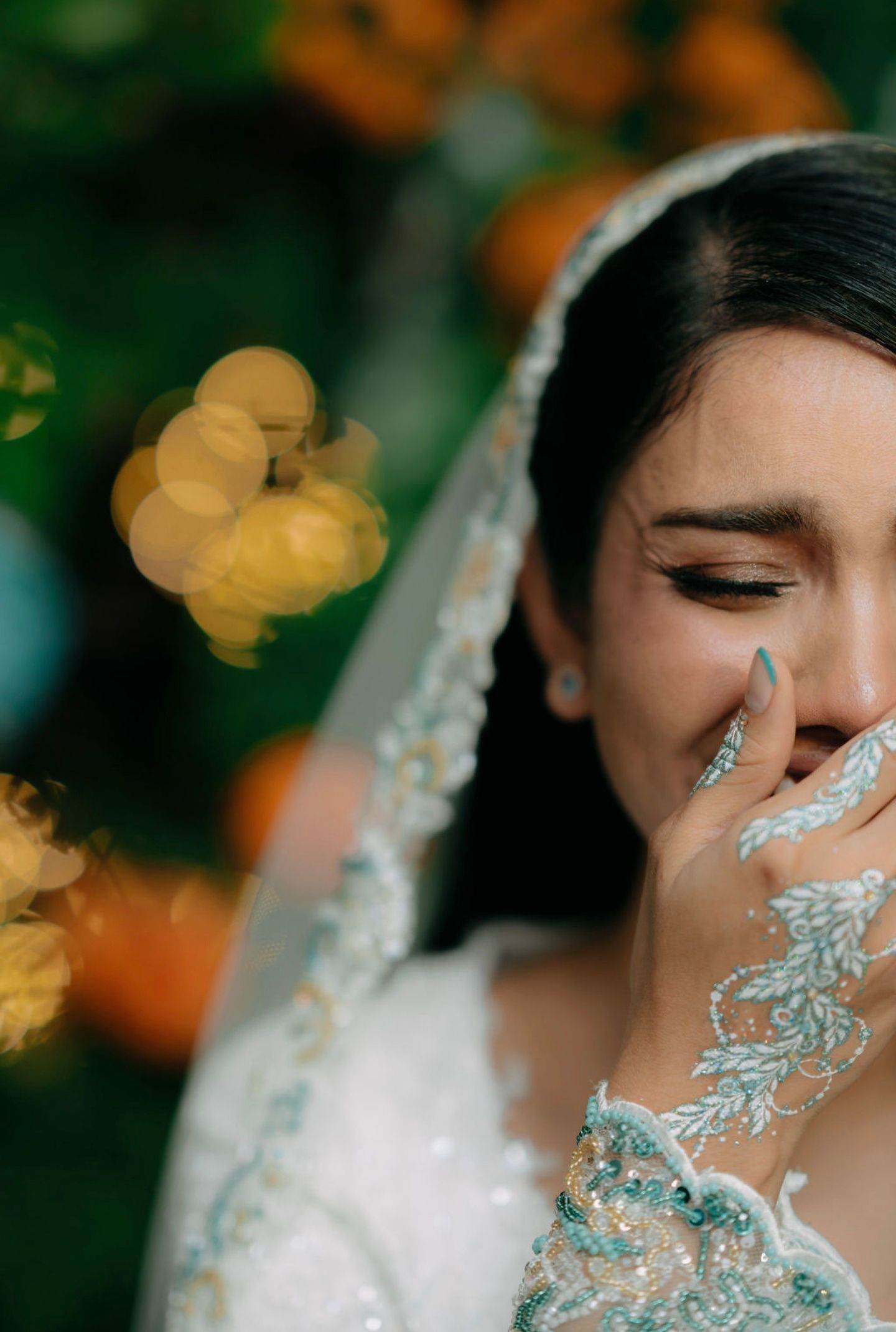 Emotional bride in white gown with teal embroidered veil covers mouth with lace-gloved hand outdoors.