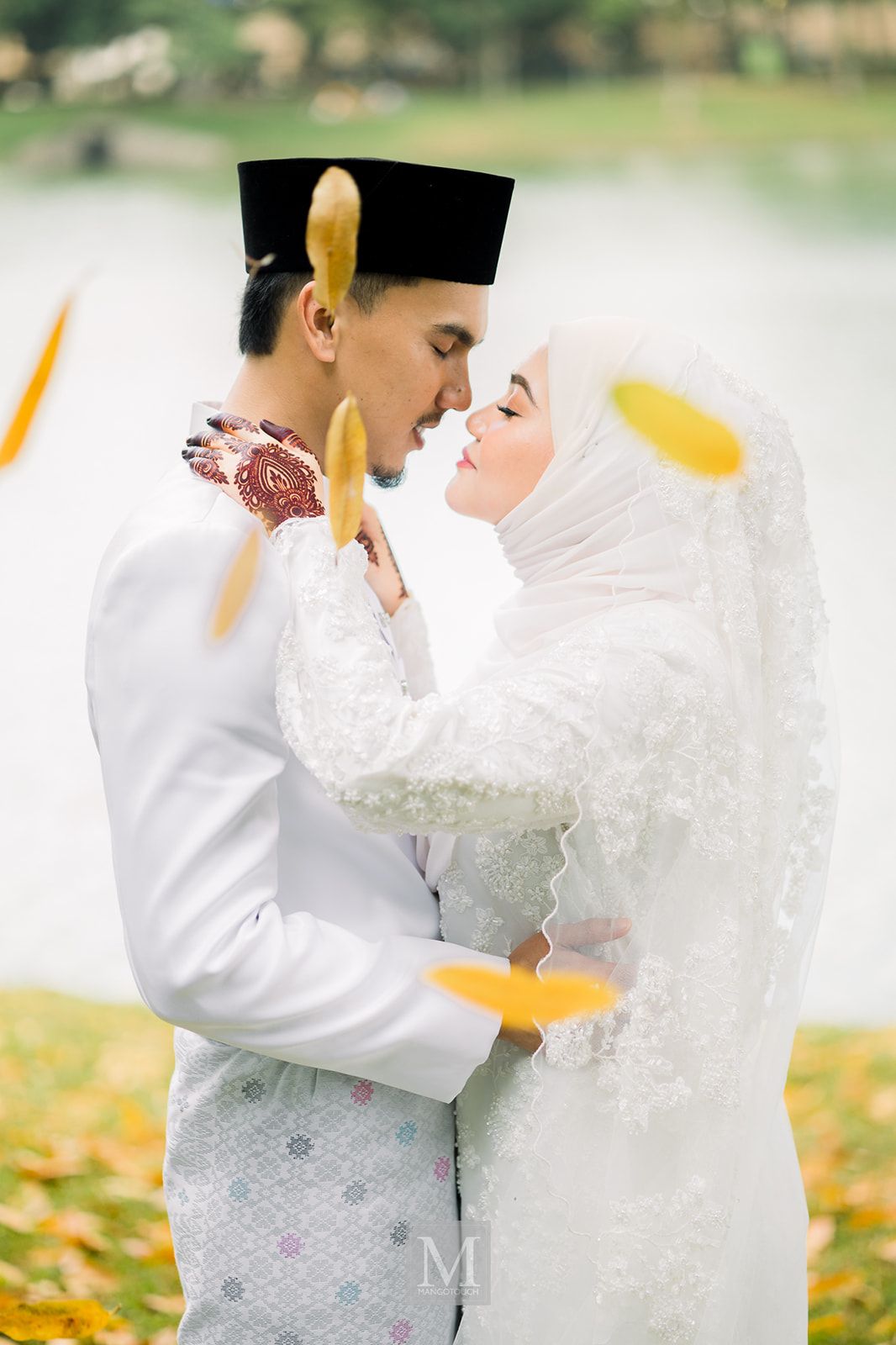 Malaysian bride and groom in white attire sharing romantic moment with falling yellow leaves.