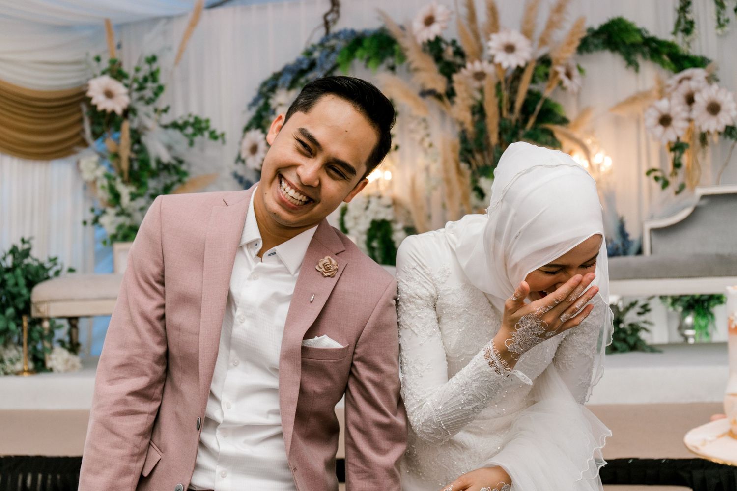 Malaysian couple laughing joyfully at their wedding reception, groom in pink suit, bride in white hijab and gown.